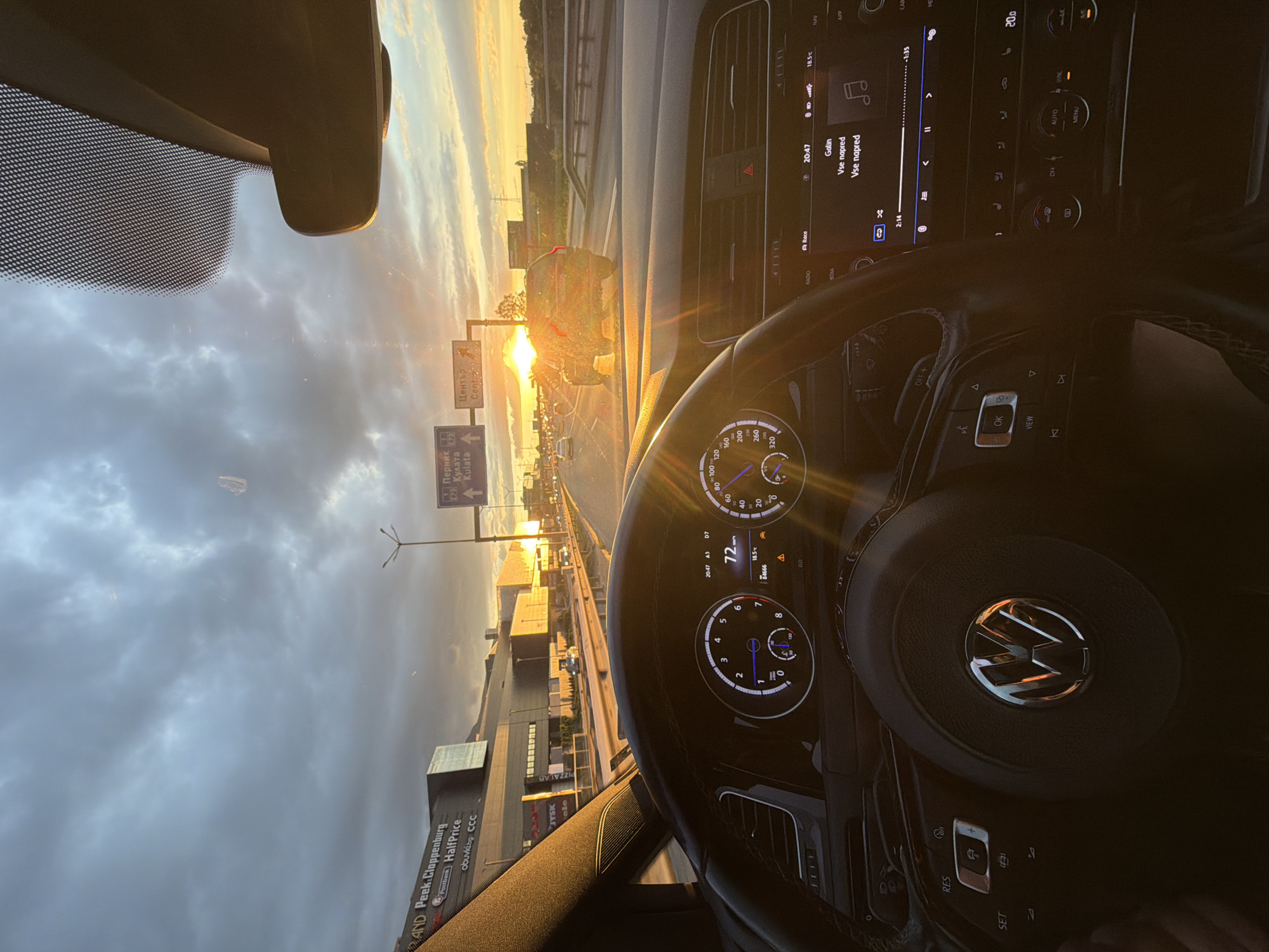 The image is taken from the driver's perspective inside a car, looking out at a highway at sunset. The car's dashboard and steering wheel are visible in the foreground, with a Volkswagen logo on the steering wheel. The digital speedometer shows a speed of 72 km/h, and the exterior temperature is 18.5°C. The car's infotainment system is displaying song information, with the artist "Galin" and the song title "Vse napred".
In the background, there is a highway with several cars, including a dark SUV driving in the right lane. The sun is setting, casting a warm glow over the scene and creating lens flare on the windshield and dashboard. On the left side of the highway, there is a large building complex with signage that includes "Peek.Cloppenburg", "Postbank", and "HalfPrice".
Overhead, there are road signs indicating directions to "Pernik", "Kulata", and "Center". These signs suggest the location is on the outskirts of Sofia, Bulgaria, likely on the A3 highway or a major road leading out of the city. The sky is filled with dramatic clouds, illuminated by the setting sun. The overall scene depicts a person driving on a highway during sunset.