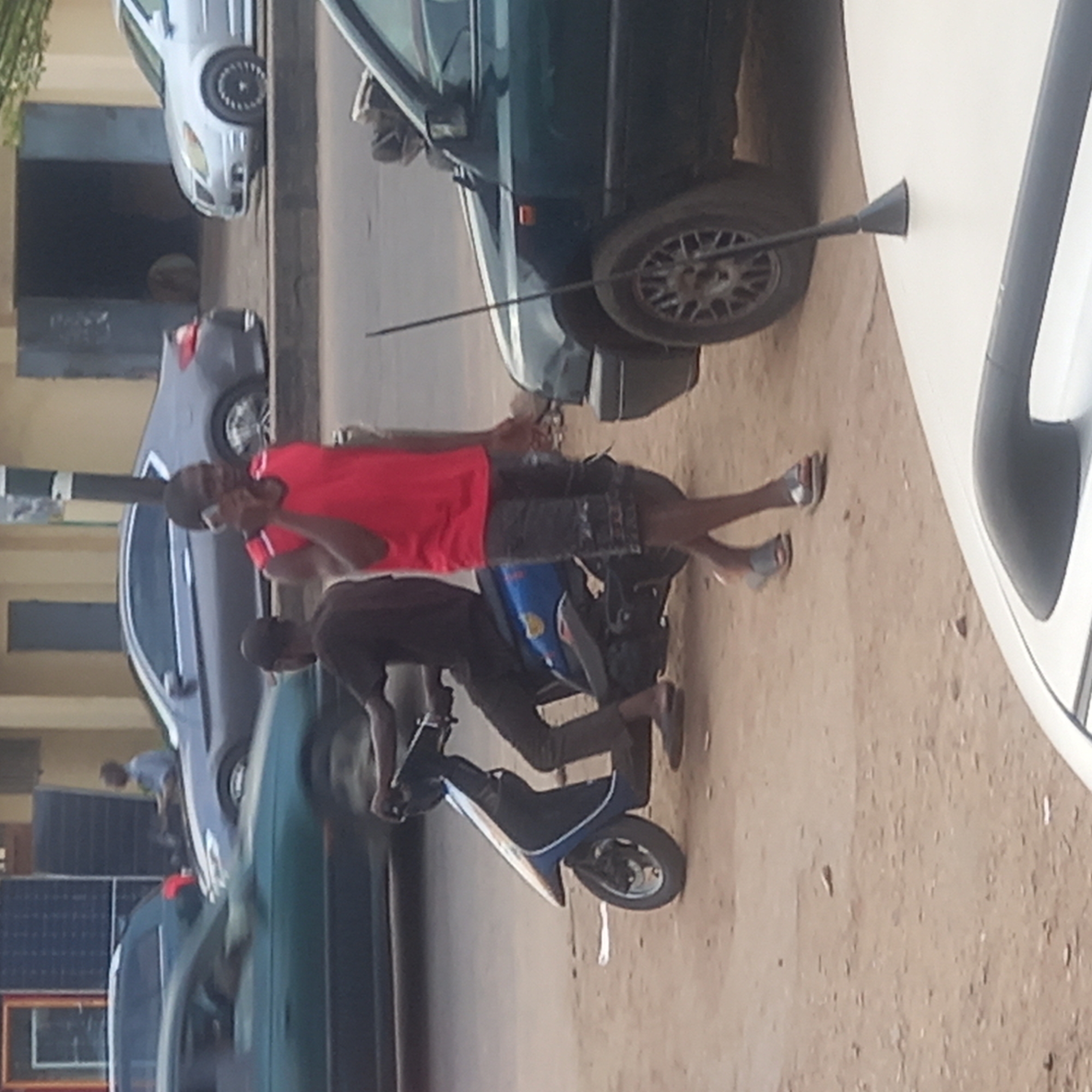 The image depicts a street scene in Dadi Riba, Nigeria. A man in a red shirt and patterned shorts stands on a scooter while talking on a cell phone. Another person is seated on the scooter, presumably operating it. Cars are parked along the street. A long, thin object resembling a pole is leaning against one of the cars. The street surface appears to be dirt or sand.