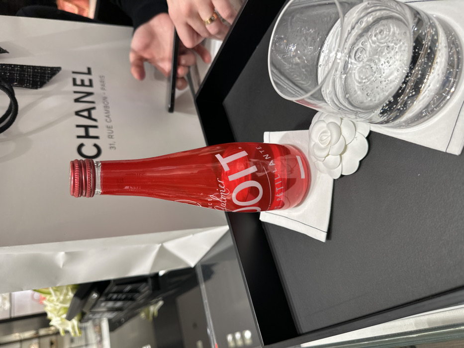 The image captures a moment within a luxury shopping environment, specifically a Chanel boutique in Paris, France. The scene is illuminated by bright indoor lighting, suggesting it's daytime.
In the foreground, resting on a sleek black tray, is a slender red glass bottle containing a reddish beverage. Its label prominently displays "P.O.T.T." in white letters, with smaller text below reading "ice Dalmier PÉTILLANTE". The bottle has a red screw cap. Beside it, a clear glass filled with sparkling water, indicated by visible bubbles, is also present. Both the bottle and the glass are placed on elegant white, camellia-shaped coasters, a distinct design element associated with Chanel.
Behind these refreshments, a white Chanel shopping bag is clearly visible. The bag features the iconic "CHANEL" logo in black, along with the address "31, RUE CAMBON - PARIS", which unequivocally places the setting. In the upper left background, two hands belonging to a person are visible. One hand wears a gold ring and holds a smartphone, suggesting the person might be browsing, taking a photo, or engaging with their device while taking a break.
The overall image conveys a sense of indulgence and luxury, characteristic of a high-end shopping experience at the famous Parisian fashion house.