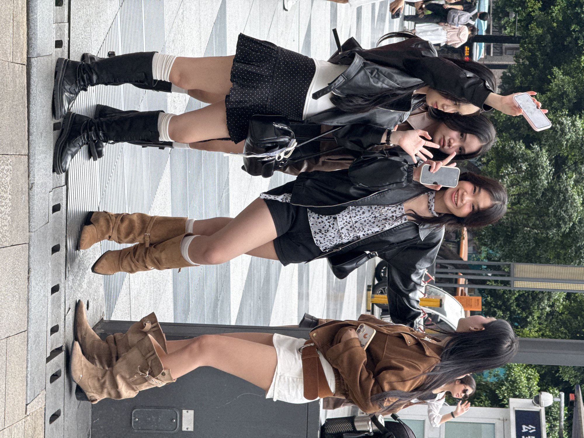 Three women are standing on a patterned walkway in Yuzhong District, China, apparently posing for a photo. The walkway is made of gray and white tiles and runs along the edge of a street where people and cars can be seen in the background. The women are standing close together, with two of them holding up their phones.
The woman at the bottom of the image is wearing a brown jacket, white shorts, and light brown boots. The woman in the middle is wearing a black jacket, patterned top, black shorts, and light brown boots with white socks. The woman at the top is wearing a black jacket, a white top, a black polka dot skirt, black boots, and white socks. The overall setting appears to be an urban, possibly commercial, area.