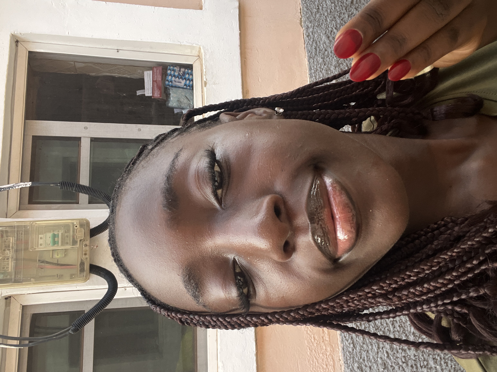 A close-up shot of a woman with dark skin and brown braided hair. She has glossy lips and red nail polish. The background includes a window and an electrical meter. The specific location within Abuja, Nigeria, is not determinable from the image.