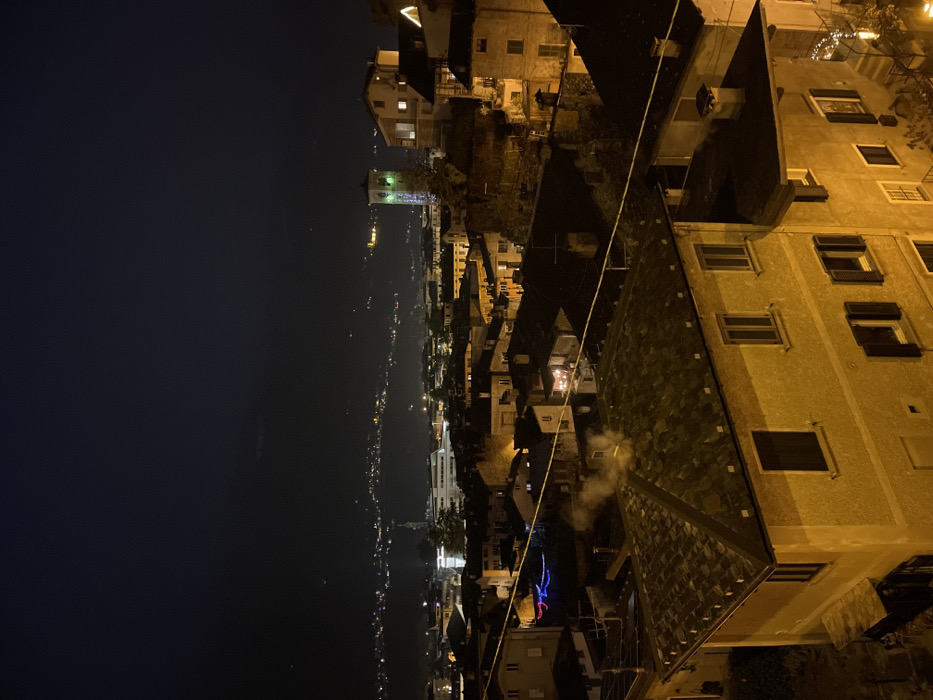The image, when properly oriented, presents a nighttime panoramic view of a town or city, likely located in a valley or along a body of water, consistent with the location context of Sondrio, Italy. The sky above is dark, indicating a late hour, with a faint glow visible on the horizon suggesting distant light pollution or the remnants of twilight.
In the far distance, numerous small lights stretch horizontally, outlining the contours of what appears to be a long body of water or a distant winding road, flanked by illuminated settlements. Moving closer to the mid-ground, more densely packed buildings are visible, characterized by warm yellow lights emanating from windows. A prominent, taller building, possibly a church tower, stands out with its own distinct lighting. Decorative, multi-colored lights (blue, purple, red) are strung on a building or structure in this middle section, adding a festive touch.
The immediate foreground features the rooftops and upper stories of several buildings. One large building dominates the right side, showing a textured, dark-tiled roof and multiple windows, some of which are illuminated. A faint plume of white smoke or vapor is seen rising from a vent or chimney on this roof, suggesting indoor warmth or activity. Another building in the center foreground also has a visible roof. A thin wire or cable runs diagonally across the scene, from the upper right to the mid-left. No people are visible, and no discernible text appears in the image. The scene conveys a quiet, serene atmosphere of a town settled into the night.