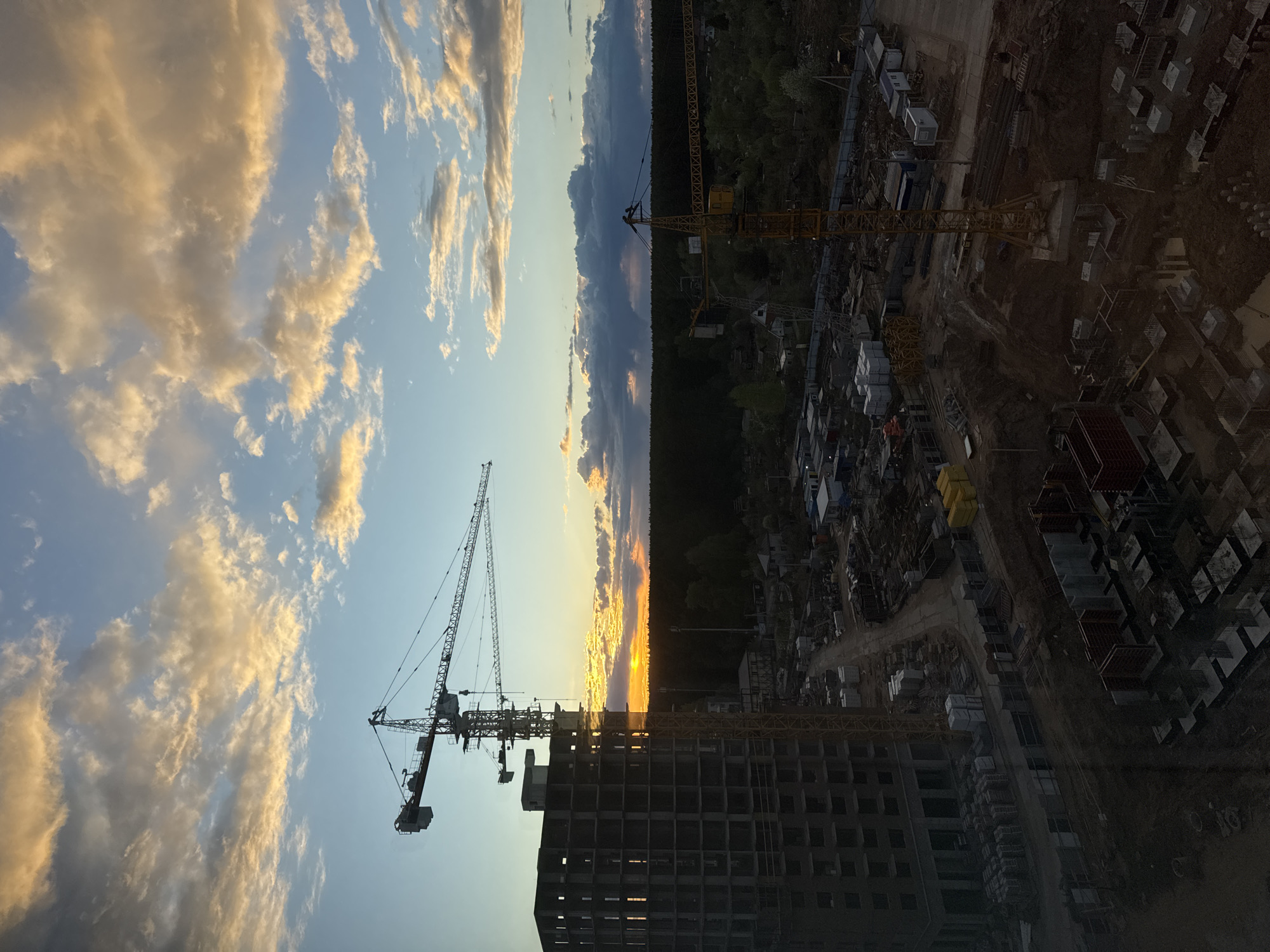 The image depicts a construction site at dusk or dawn in Izhevsk, Russia. A high-rise building under construction dominates the foreground, with multiple cranes positioned around it. The sky features a mix of blue and orange hues, with clouds scattered across the horizon. Beyond the construction site, a dense forest borders a residential area. The overall scene suggests ongoing urban development within Izhevsk.