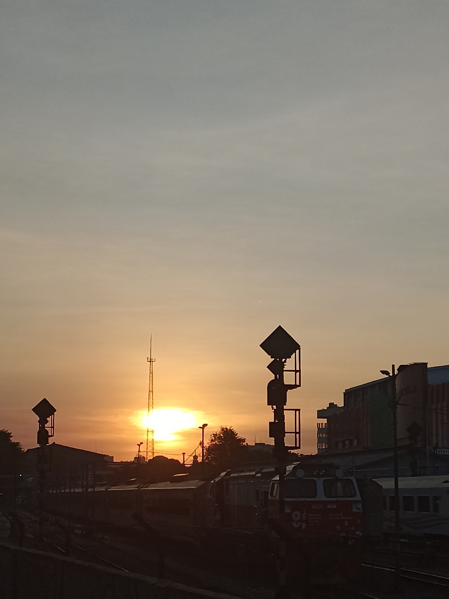 The image captures a sunrise scene at a train station in Surabaya, Indonesia. The sky is a gradient of light blue to pale orange, with the sun a bright white orb rising behind a tall communication tower. In the foreground, railway tracks stretch into the distance, flanked by several trains. A prominent red and white train engine is visible on the right side of the frame. Silhouetted against the sky are signal posts, buildings, and trees. The overall atmosphere is serene, suggesting the early morning hours at a busy transportation hub.