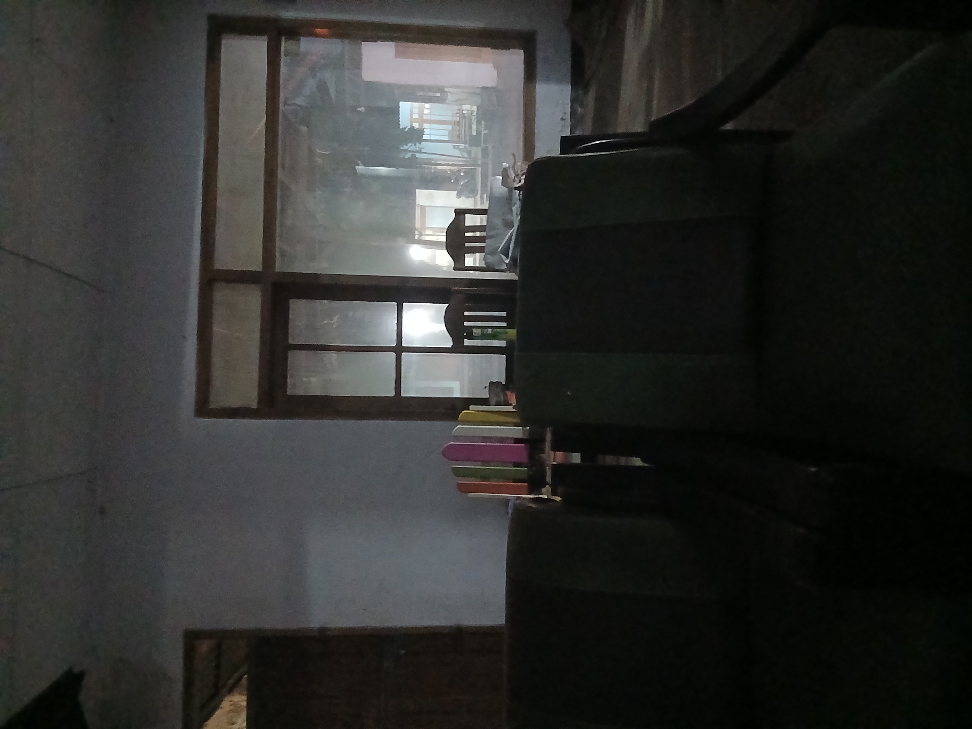The image captures an indoor scene with two dark upholstered sofas in the foreground, facing away from the viewer. Behind the sofas, a large window with wooden framing dominates the back wall, revealing a dimly lit outdoor area. The outdoor view shows what appears to be a street or pathway with buildings, a parked motorcycle, and some outdoor seating.
To the left of the window, a wooden door is visible, partially obscured by the wall. In front of the window, two wooden chairs with slatted backs are positioned. On a surface to the left of the chairs, a colorful, arched object resembling a stack of books or decorative items is present, with hues of pink, green, orange, and yellow. The room itself has plain walls, and the lighting is low, suggesting it may be evening or nighttime. The overall impression is of a domestic or communal living space, possibly a waiting area or lounge, with a view of an outdoor environment. The location is Kademangan Kulon, Indonesia.