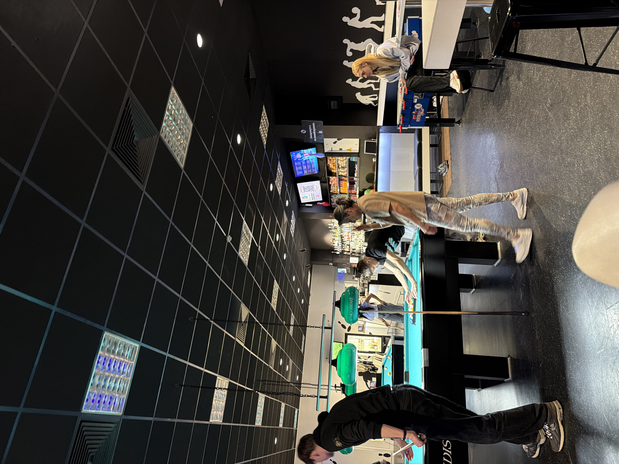 This image captures an indoor scene within "Bowling Paradise," a recreational establishment located in Paradise Center, Sofia, Bulgaria. The environment is characterized by a dark, grid-patterned ceiling fitted with square fluorescent lights, some emitting blue and purple hues, along with recessed spotlights and HVAC vents.
In the foreground-left, a male individual in a black hoodie and dark pants stands, holding a pool cue with his left hand, next to a teal-felted pool table. His back is mostly to the viewer. In the mid-ground-right, another male, wearing a dark t-shirt and light pants, leans over a separate teal-felted pool table, in the process of racking or arranging billiard balls. Between these two tables, a female with long hair, wearing a light brown top and patterned leggings, walks from left to right. Overhead, two green-shaded industrial lamps hang from chains, illuminating the pool tables. The word "SISSDIS" is visible on the side of one lamp fixture.
To the right, a blonde female in a light grey hoodie and dark pants is seated on a black high stool at a white high-top table, positioned next to a blue foosball table. The background features a bar area with multiple shelves stocked with liquor bottles, visible television screens, and a counter. A white sign with Bulgarian text "Задължително е на самообслужване" (Self-service is mandatory) is affixed to the wall near the bar. The wall also displays white silhouette art depicting a sequence of figures in various poses, including athletic activities like bowling and basketball. The floor throughout the venue is a dark, possibly industrial-style surface with visible wear patterns. Additional individuals can be seen in the deeper background interacting with other pool tables.