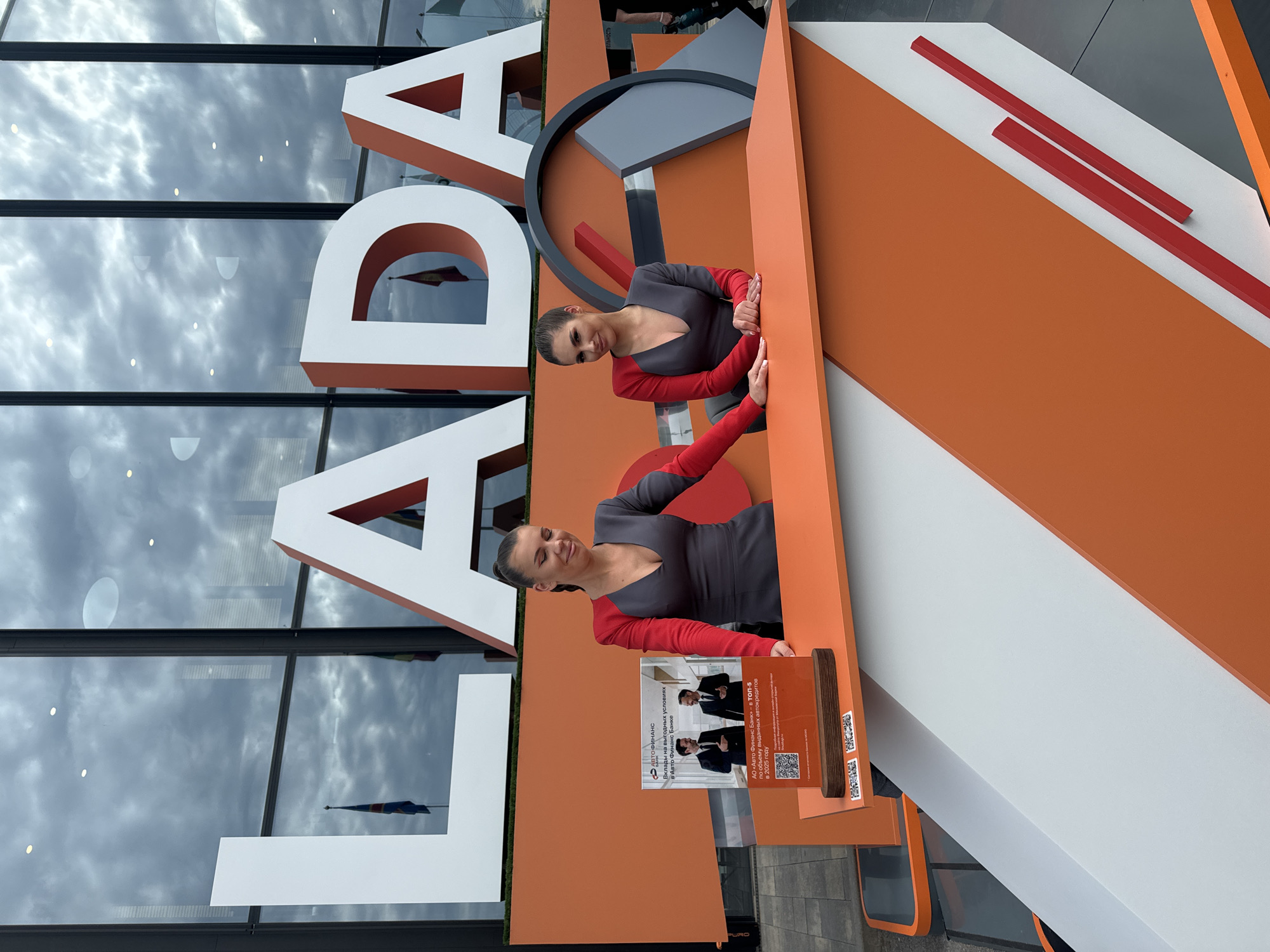 This media file captures two women standing beside a large, prominent "LADA" sign, likely at an event or dealership in Shushary, Russia. The image is taken outdoors during the daytime, with reflections of a cloudy sky visible in the glass building behind the sign.
The two women, possibly models or promoters, are positioned on the right side of the large sign. They are dressed in identical outfits consisting of a dark grey top with a deep V-neck and contrasting bright red long sleeves. Their dark hair is pulled back. One woman looks directly at the camera with a smile, while the other looks slightly away. They are both leaning on the orange part of a large, geometric display.
The central subject is a colossal "LADA" sign, featuring white dimensional letters with orange sides, set against a backdrop of orange, white, and dark grey geometric shapes. To the right of the women, there is an information stand made of wood and clear acrylic.
Visible text on the information stand includes:
- "АВТО ФИНАНС" (Auto Finance)
- "Вклады на выгодных условиях в Авто Финанс Банке" (Deposits on favorable terms at Auto Finance Bank)
- "АО «Авто Финанс Банк» в ТОП-5 по объему выданных автокредитов в 2025 году" (JSC "Auto Finance Bank" in TOP-5 by volume of issued car loans in 2025).
Below this text, two QR codes are visible.
In the background, a modern building with large reflective glass windows stretches across the scene. These windows reflect a cloudy sky and a distant flag, providing a sense of the outdoor environment. The overall impression is one of a promotional or corporate event related to the LADA brand and associated financial services.