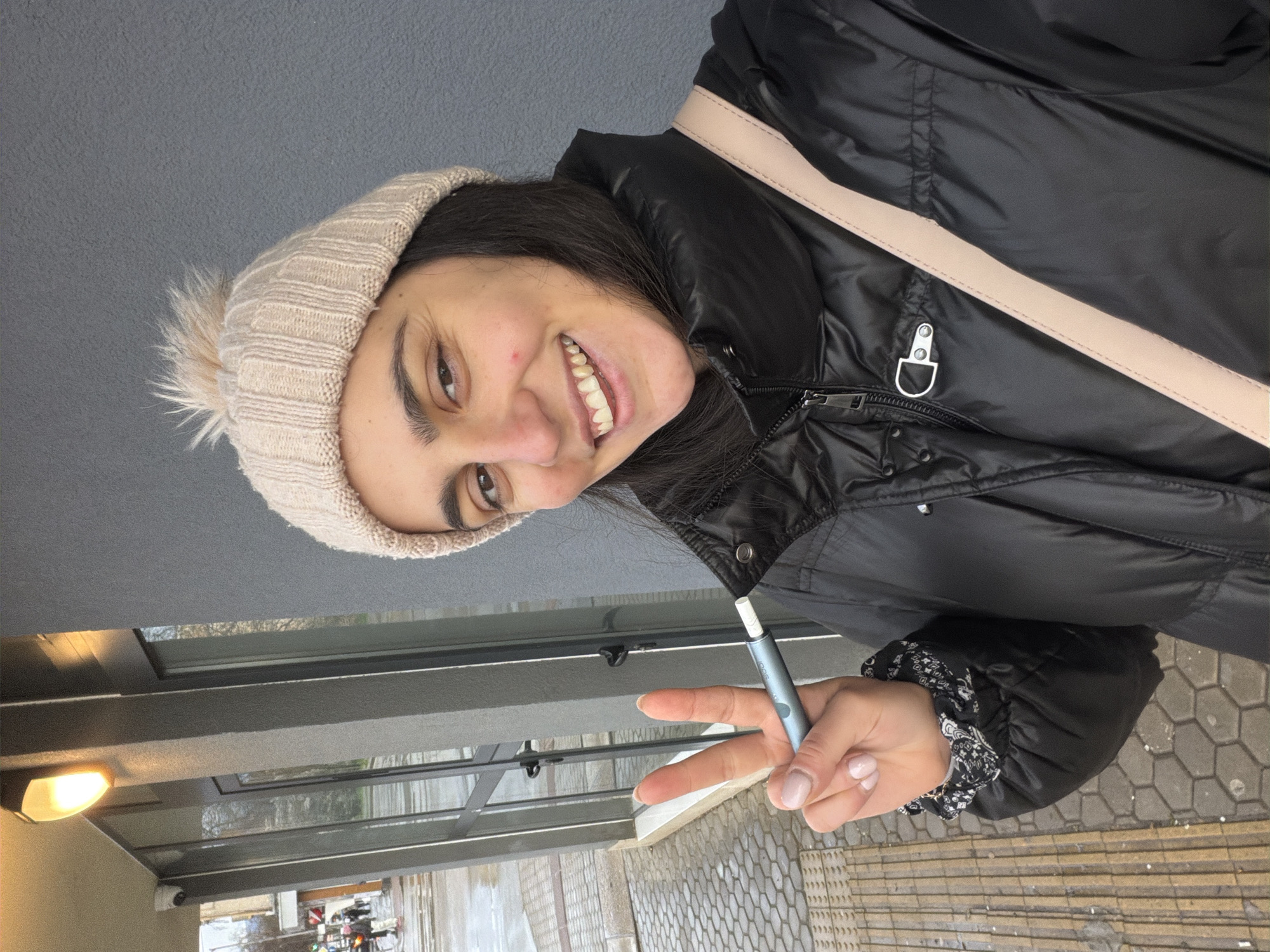 A woman is shown holding a smoking device. She is wearing a beige knit hat with a pom-pom, and a black puffer jacket. A light-colored strap crosses diagonally across her chest. She is making a peace sign with the hand holding the smoking device. The background appears to be an outdoor urban setting, possibly a sidewalk or pedestrian area, with a glass-covered structure or bus stop behind her. Based on visual clues, this image was likely taken in Sofia, Bulgaria.