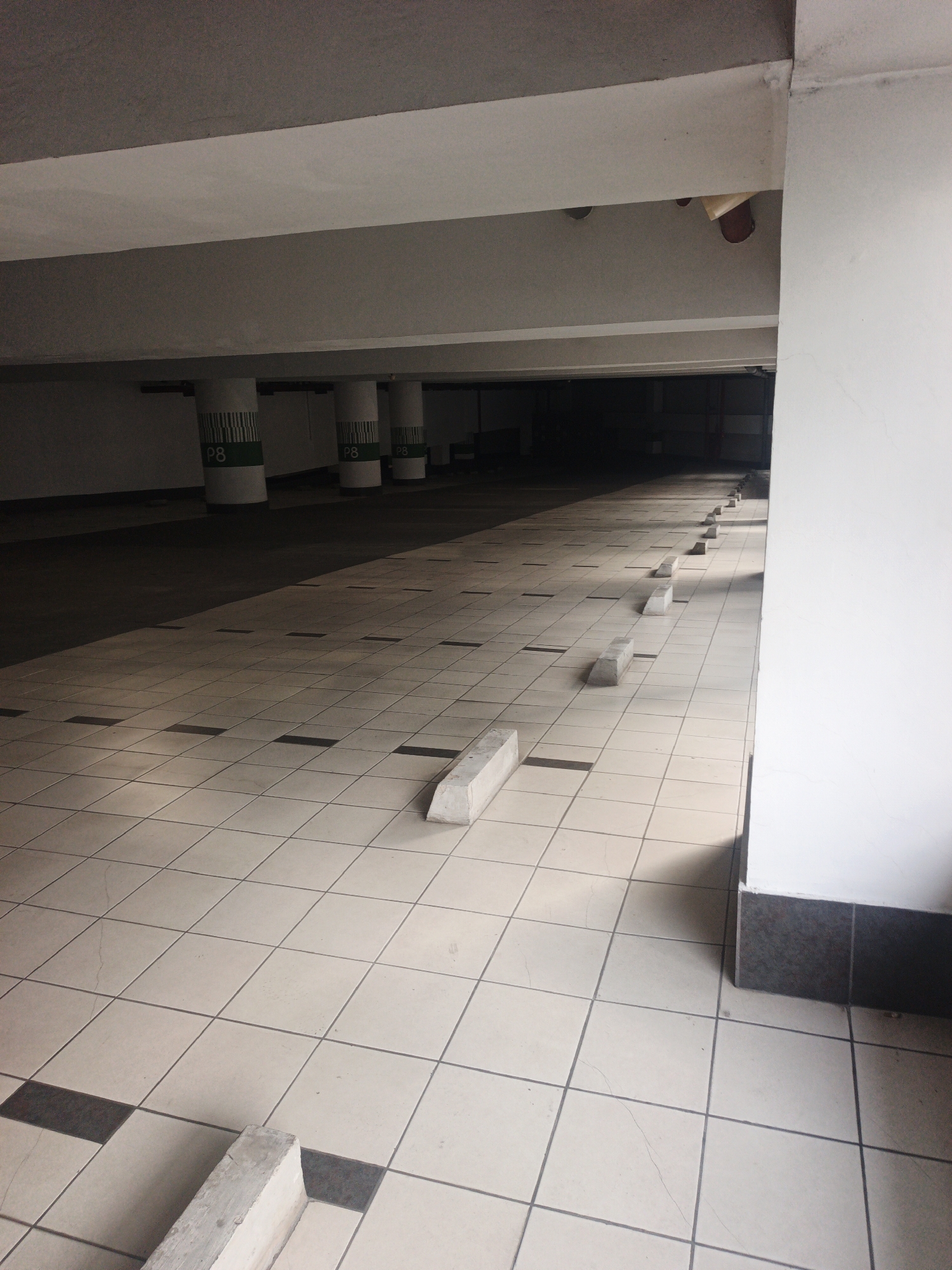 This is a photograph taken from a low angle inside a dimly lit, tiled indoor parking garage. The floor is covered in light-colored square tiles with darker square accents, laid out in a grid pattern. Several concrete parking stops are visible on the floor, marking individual parking spaces.
In the background, cylindrical concrete pillars are arranged in a row. These pillars are painted white with green bands near the top, on which the text "P8" is visible in black lettering. The ceiling of the parking garage is visible, appearing to be constructed of concrete beams and panels. The lighting is subdued, casting long shadows across the tiled floor. The overall impression is one of an empty, utilitarian space.
The location is identified as a parking garage in South Jakarta, Indonesia.