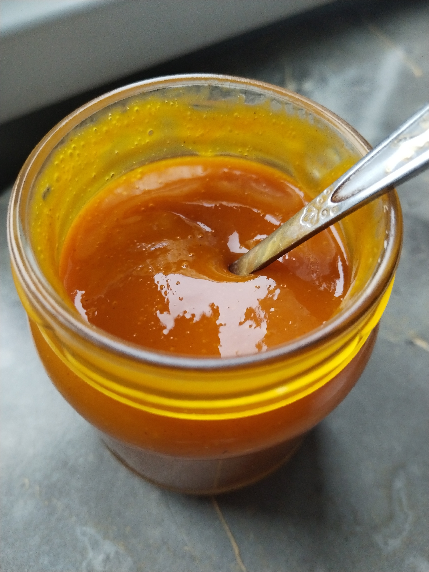 A clear glass jar contains a viscous, orange-brown substance, consistent with jam or sauce, on a dark gray, mottled countertop. A silver-colored spoon is partially submerged within the substance, resting against the inner rim of the jar. Reflections of ambient light are visible on the surface of the substance and on the spoon. Residue from the substance adheres to the interior upper walls of the jar. The background is a blurred field of gray and black tones. This scene is located in Бараўлянскі сельскі Савет, Belarus.