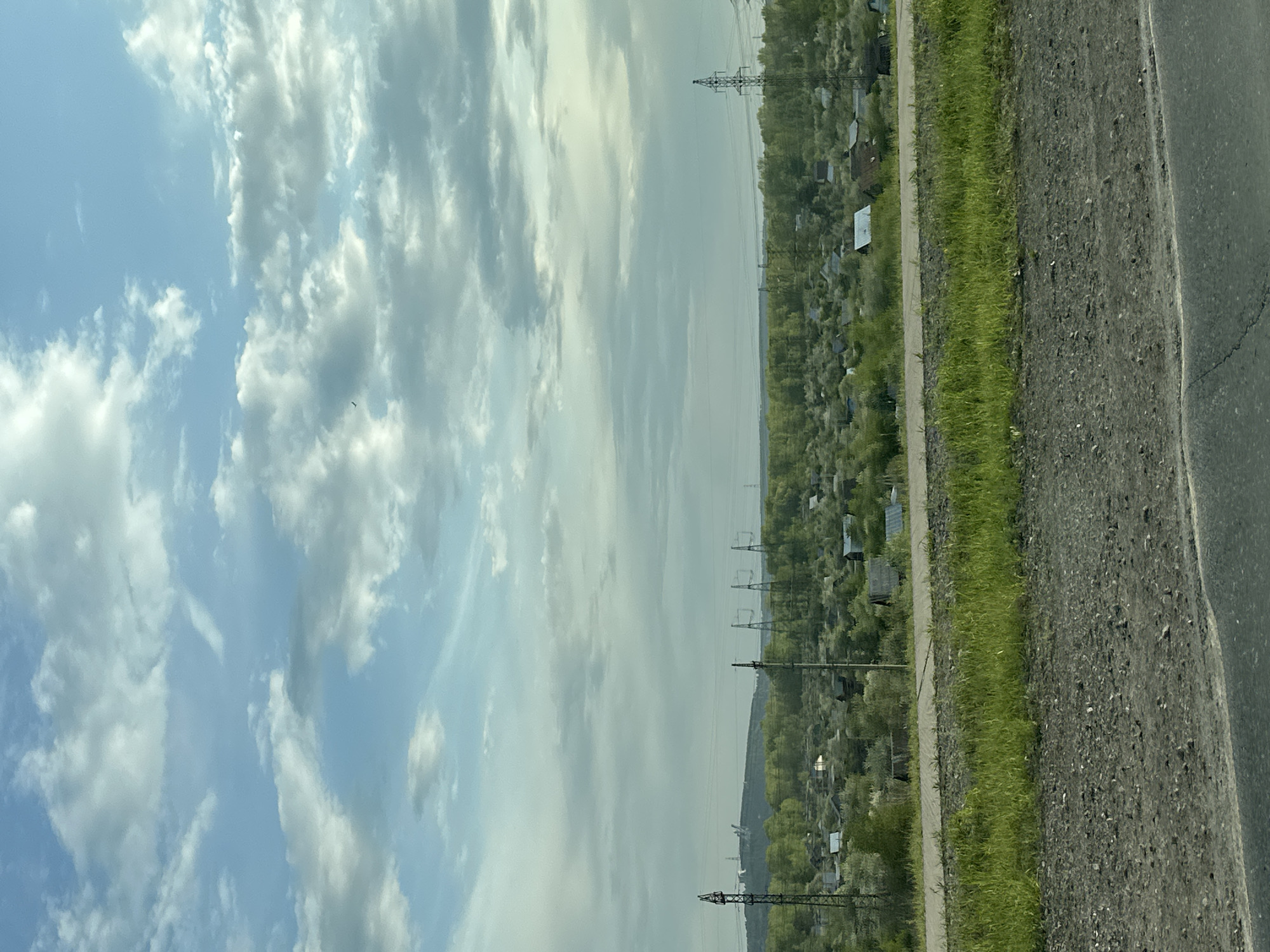 The image presents a roadside view in the city of Chaikovsky, Russia. The composition is divided horizontally, with the sky occupying the left portion and the landscape on the right. The sky features scattered clouds with varying densities against a backdrop of blue. The roadside scene includes a stretch of road, a grassy verge, and a dense area of vegetation interspersed with buildings, possibly a residential or allotment area. A series of electricity pylons are visible running through the landscape, suggesting an industrial or semi-rural setting. The road surface appears to be asphalt, showing signs of wear. There are no discernible people or specific actions visible in the image.