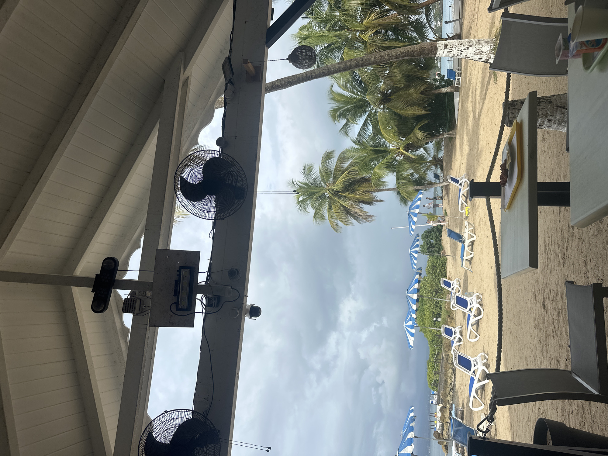 The image shows an outdoor scene at a beach in Le Gosier, France. The view is from under a partially covered structure, likely a restaurant or bar.
**Environment:** The beach is sandy with palm trees and blue and white striped beach umbrellas. Several beach chairs are visible. The sky is overcast with grey clouds.
**Objects:** Two black fans are attached to the ceiling of the structure. A spotlight, a lighting box, and a security camera are also mounted on the ceiling. Tables and chairs are visible on the right side of the frame. A food tray with brown objects is on one of the tables.
**People:** No distinct people are visible.
**Actions:** No actions are directly visible.
**Interactions:** The interaction between the covered structure and the outdoor environment is evident.