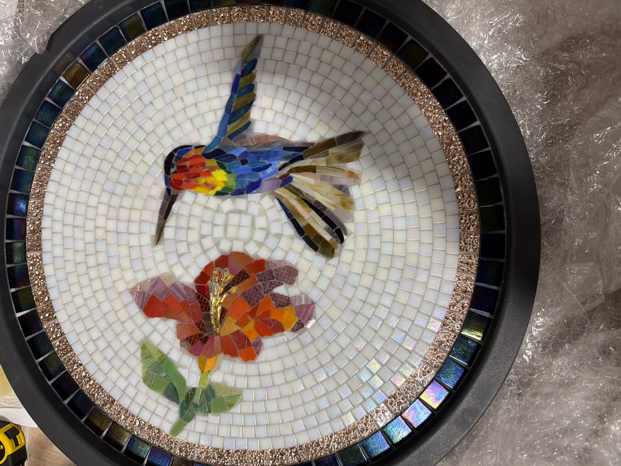 A circular mosaic art piece featuring a hummingbird and a flower is displayed on a white background. The mosaic is bordered by concentric rings of small white tiles, followed by a ring of textured rose gold tiles, and then a ring of iridescent square tiles in shades of blue and green. The outermost edge is a dark grey rim. The hummingbird is rendered in vibrant colors, with blues, reds, yellows, and greens forming its body and wings. The flower is depicted in shades of orange, red, and purple, with green leaves. The entire mosaic is set within a dark grey circular frame, and appears to be placed on a surface covered with bubble wrap. The location is Lower Southampton Township, United States of America.