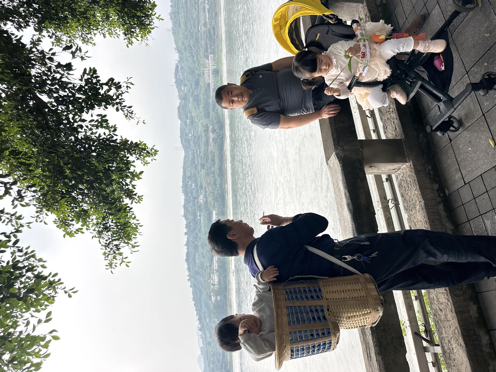 The image shows an outdoor scene in Jiangjin, China, likely taken along the Yangtze River. A father and daughter are seated by the river's edge next to a stroller with a child. The adult male is wearing a grey collared shirt. A stroller is positioned next to them with a young child seated inside wearing a dress. On the other side of the walkway, another man, dressed in a dark jacket, is standing with a child in a basket strapped to his back. He is holding a cigarette in his hand. The river flows behind them, with forested hills on the opposite bank. The sky is overcast. A leafy tree branch is visible in the upper left of the frame. The ground is a paved walkway with a metal railing visible between the two men.