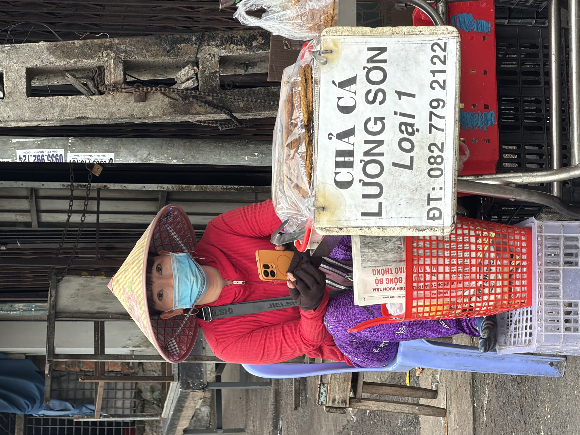 A woman wearing a conical hat and a blue face mask sits on a blue plastic chair in front of a stall in Nha Trang, Vietnam. She is wearing a red long-sleeved shirt and purple pants with a leopard print pattern. She is holding an orange smartphone in her right hand, which is gloved. In front of her is a red plastic basket filled with newspapers. A sign in front of the stall reads "CHẢ CÁ LƯƠNG SƠN Loại 1 ĐT: 082 779 2122". Behind the sign, packages of fried fish cakes are visible. The stall is set up on a sidewalk with a metal grate and other stalls visible in the background. A locked chain is attached to a pole to the right of the woman. The overall scene depicts a street vendor in Vietnam.