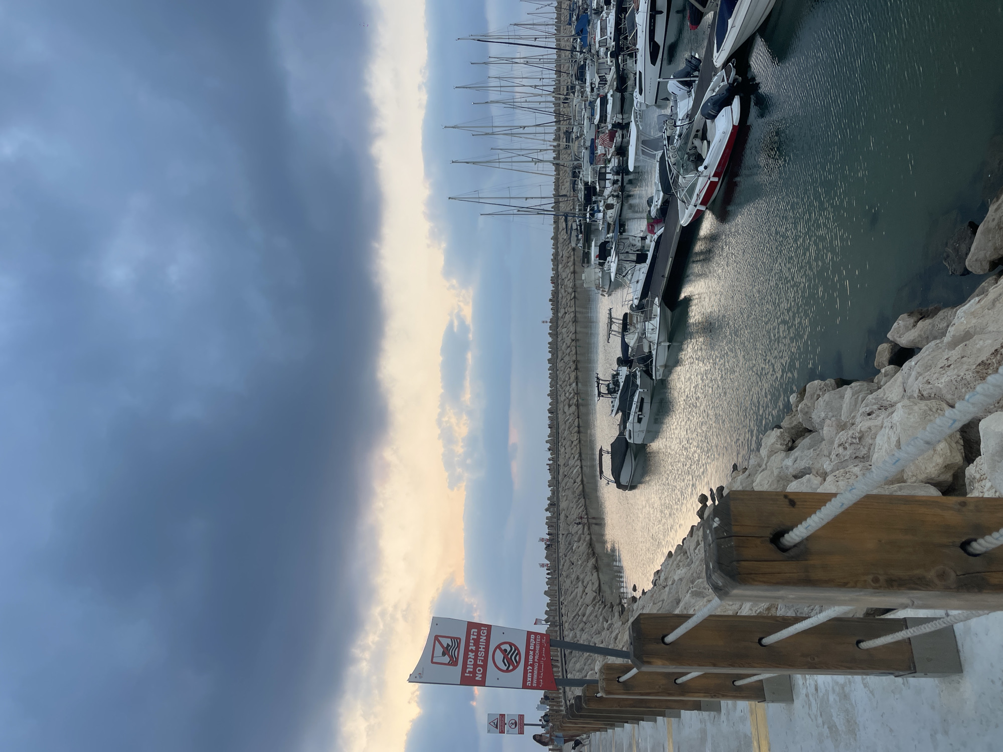 The image depicts a marina in Herzliya, Israel, with a dramatic sky filled with dark, swirling clouds. In the foreground, a paved walkway lines the edge of the marina, bordered by a wooden fence with thick ropes. A sign prominently displays "NO FISHING!" in English and Hebrew, with an accompanying symbol of a crossed-out fishing rod.
The marina itself is filled with numerous boats of various sizes, from small motorboats to larger sailboats, all moored along docks. The water's surface reflects the muted light of the sky and the surrounding structures. Along the far edge of the marina, a stone breakwater stretches into the distance, with people visible walking along it. The overall impression is one of a tranquil evening at the harbor, despite the foreboding clouds.