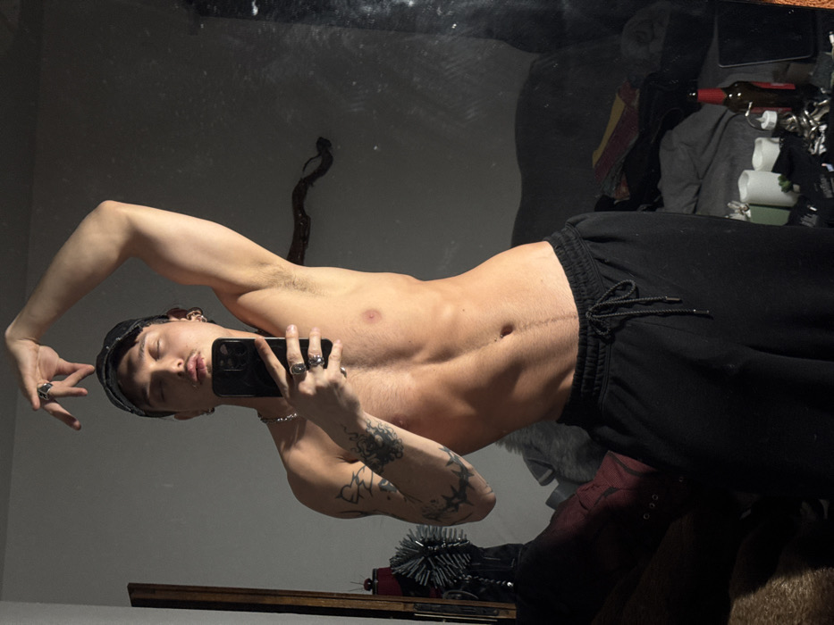A male individual is depicted in a mirror selfie, standing shirtless and wearing black drawstring sweatpants and a dark baseball cap. He possesses a lean, muscular physique with visible abdominal musculature. Tattoos are present on his left arm, consisting of an abstract design on the bicep/shoulder area and a darker, textured pattern on the forearm. He wears multiple silver-colored rings on his fingers and earrings in his left ear, along with a silver-toned chain around his neck. His eyes are closed or heavily lidded, and he exhibits light facial stubble above his lip and on his chin. His left arm is raised and bent with the hand positioned above his head, fingers forming a stylized gesture. His right arm is bent, holding a black smartphone, which is angled to capture his reflection in the mirror. His body is slightly angled and curved, emphasizing his torso.
The background, visible in the mirror's reflection, indicates an indoor environment, likely a private room. To the reflected left of the subject, a dark vertical furniture piece is partially visible, alongside a dark, bristled object and a dark red item resembling a boot or textile. The wall appears dark gray or off-white. To the reflected right, a gnarled, dark wooden branch or decorative element is affixed to the wall. Further right, a dark sofa or unmade bed with rumpled textiles is discernible. On a surface in the lower reflected right, several white cylindrical objects (possibly candles), a brown glass bottle with a red label, a white squeeze bottle, and a collection of small, reflective items are arranged among dark fabrics.
The specific location within Sofia, Bulgaria, cannot be ascertained from the visual evidence presented in the image.