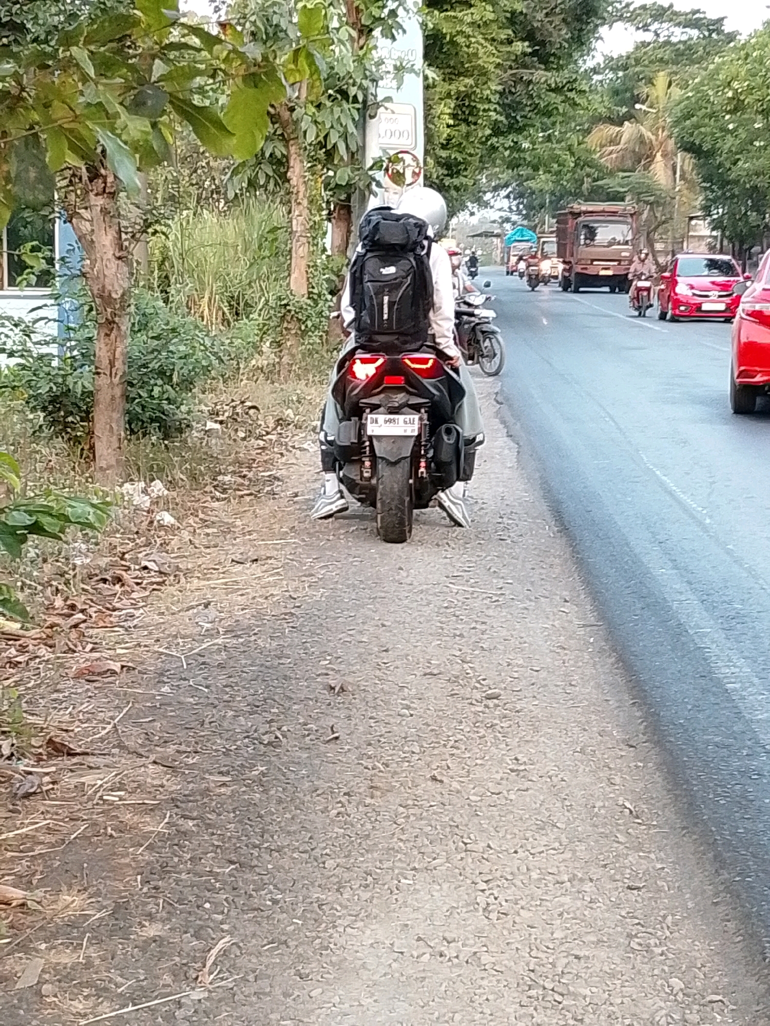 A person on a scooter with a large backpack is traveling along a road in Pasuruan, Indonesia. The scooter has a license plate that reads "DK 6981 GUU". The road appears to be a two-lane road with traffic. There are trees and vegetation on the side of the road. The scene is daytime.