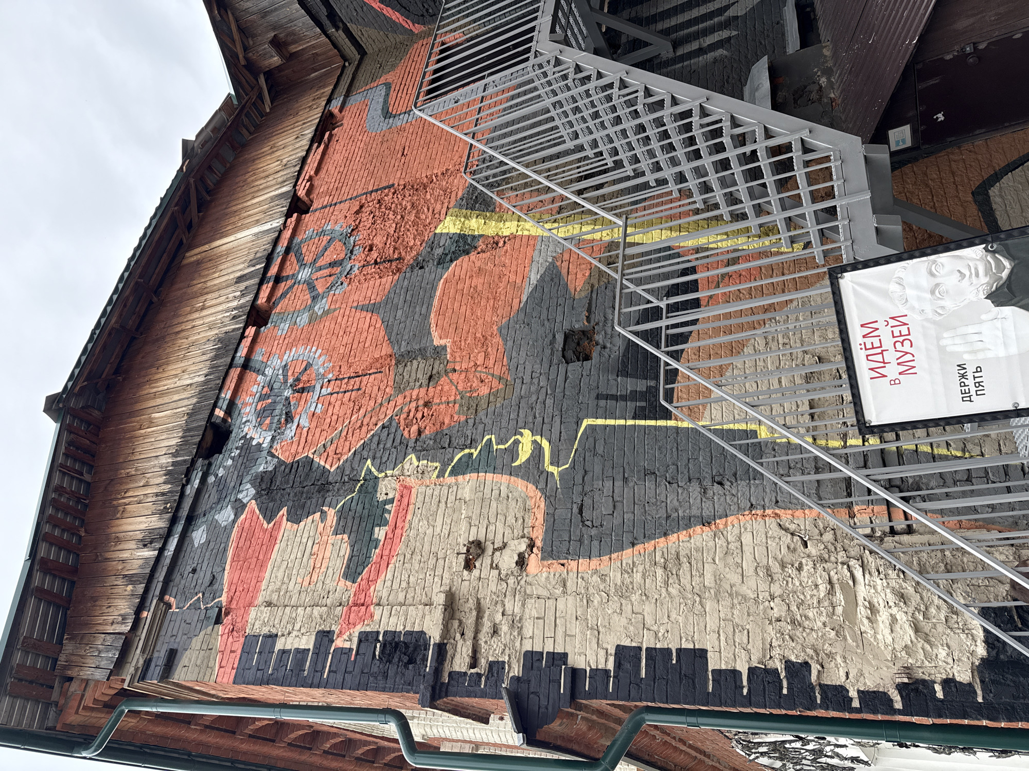 The image shows a building exterior in Votkinsk, Russia, featuring a large mural painted directly onto a brick wall. The mural appears to depict stylized industrial or mechanical elements, incorporating what seem to be wheels, abstract shapes, and a cityscape silhouette along the bottom edge. The color palette includes shades of red, gray, black, and cream.
A metal fire escape ladder is attached to the building, partially obscuring the mural. A sign hangs on the right side of the building, with text in Russian and a black-and-white image of a classical sculpture. The building itself appears to be somewhat aged, with visible wear and tear on the brickwork and mural. A wooden roof overhangs the brick wall, indicating architectural features of the building. The sky is overcast.