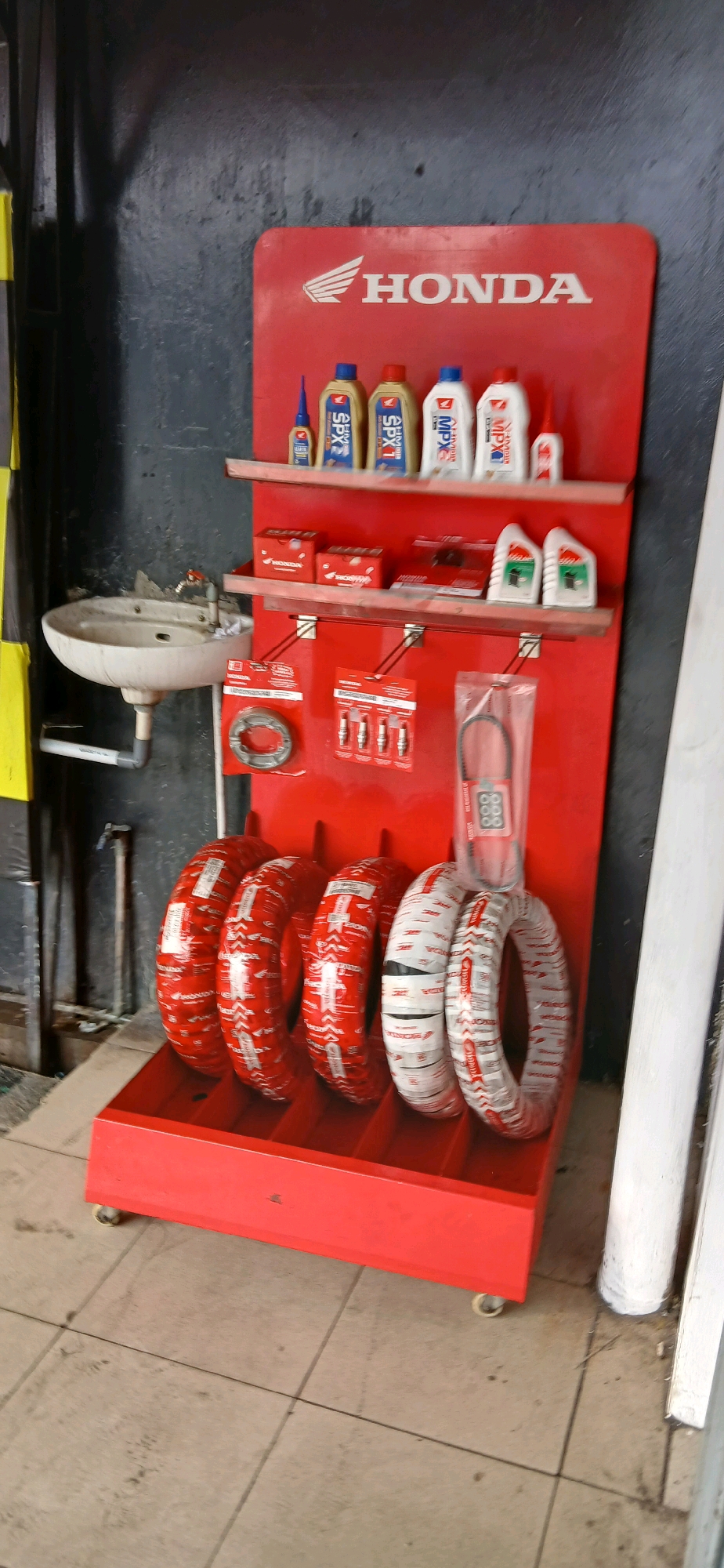 This image depicts a display rack with various automotive parts and supplies, likely in a shop or service center, located in Samarinda, Indonesia. The rack is red and features several shelves.
On the top shelf, there are multiple bottles of liquid, appearing to be oil or coolant, with labels indicating "HONDA" and "MPX," as well as other smaller bottles of what might be adhesives or lubricants.
The second shelf from the top holds several red boxes, some with the "HONDA" logo. There are also two white bottles with green and white labels, also appearing to be automotive fluids.
The third shelf from the top displays several individual parts. On the left, there's a circular metal part, possibly a brake disc or clutch component, in a plastic package. Next to it are several spark plugs, also packaged. To the right, a black belt, likely a drive belt, is visible in a clear plastic package.
The bottom of the display unit holds several tires, wrapped in red and white packaging. The packaging features the "HONDA" logo and other text.
To the left of the display rack, a white sink is mounted on the wall. A faucet is visible, and pipes connect the sink to the wall and floor. The background wall is dark and appears to be textured. The flooring is tiled with a neutral color.
The overall impression is a retail or service environment focused on Honda motorcycles or vehicles, situated within an urban setting in Samarinda.