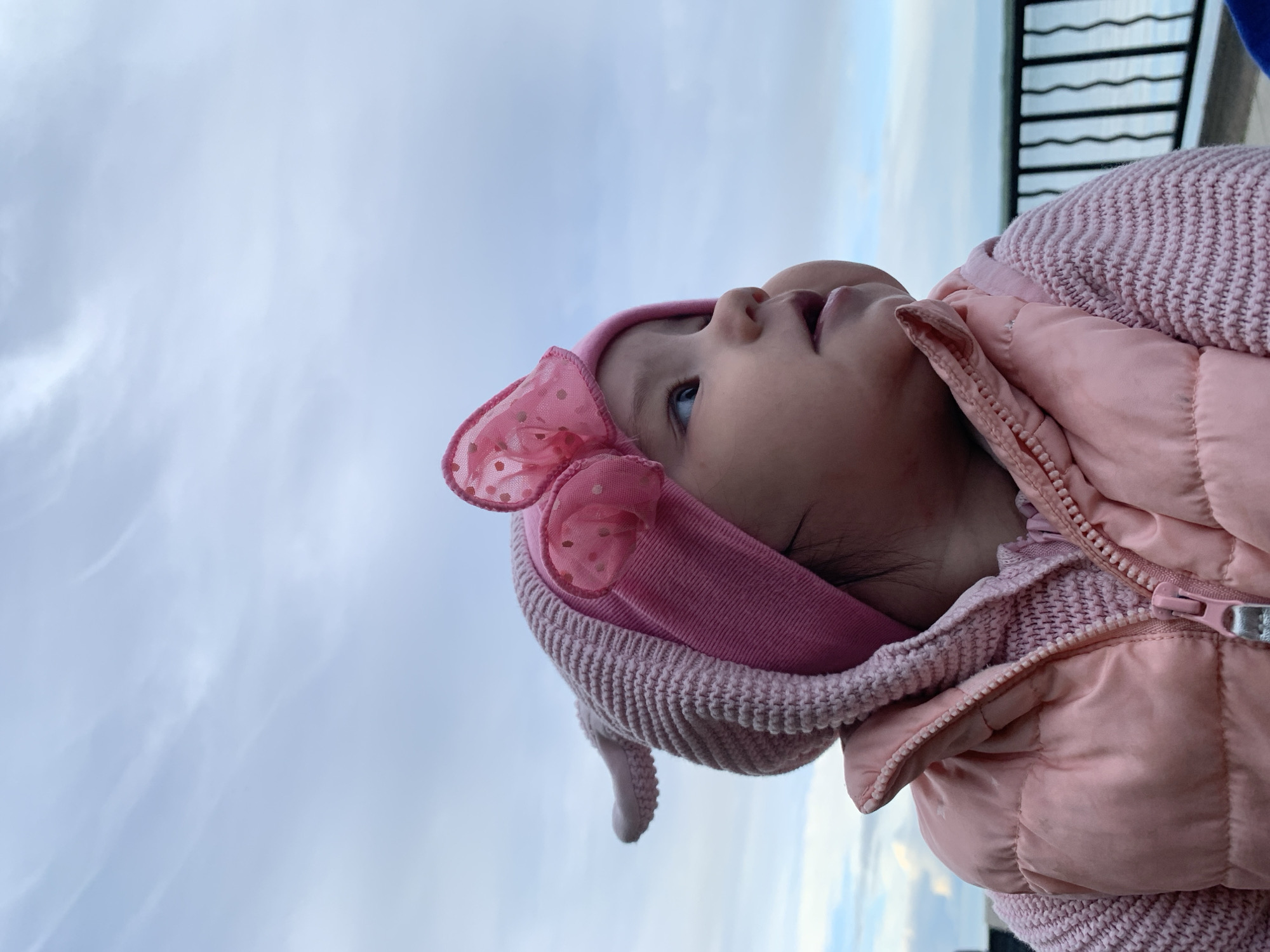 A baby in a pink jacket and hat with a bow is looking up at the sky. The background is a cloudy sky and a black metal fence. The location is Chaikovsky, Russia.