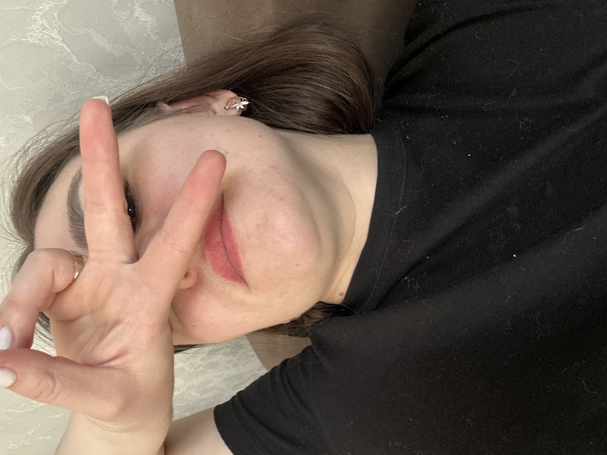 A close-up portrait shows a female subject located indoors in Chaikovsky, Russia. The subject's face is partially obscured by her right hand, held in a horizontal "V" gesture, covering her eyes. Her left eye, visible beneath her fingers, is brown. She has shoulder-length, medium-brown hair and fair skin, with visible pores and minor blemishes. Her lips are coated with pink lipstick. She wears a black long-sleeved garment, which exhibits lint and fibers. A gold-toned ring is present on the ring finger of her right hand, and a gold-toned earring, designed with a floral or butterfly motif featuring a clear central stone, is visible in her right earlobe. Her fingernails are manicured with white polish. The background features a light-colored, subtly patterned wall on the left and a plain brown upholstered surface, consistent with furniture, on the right.