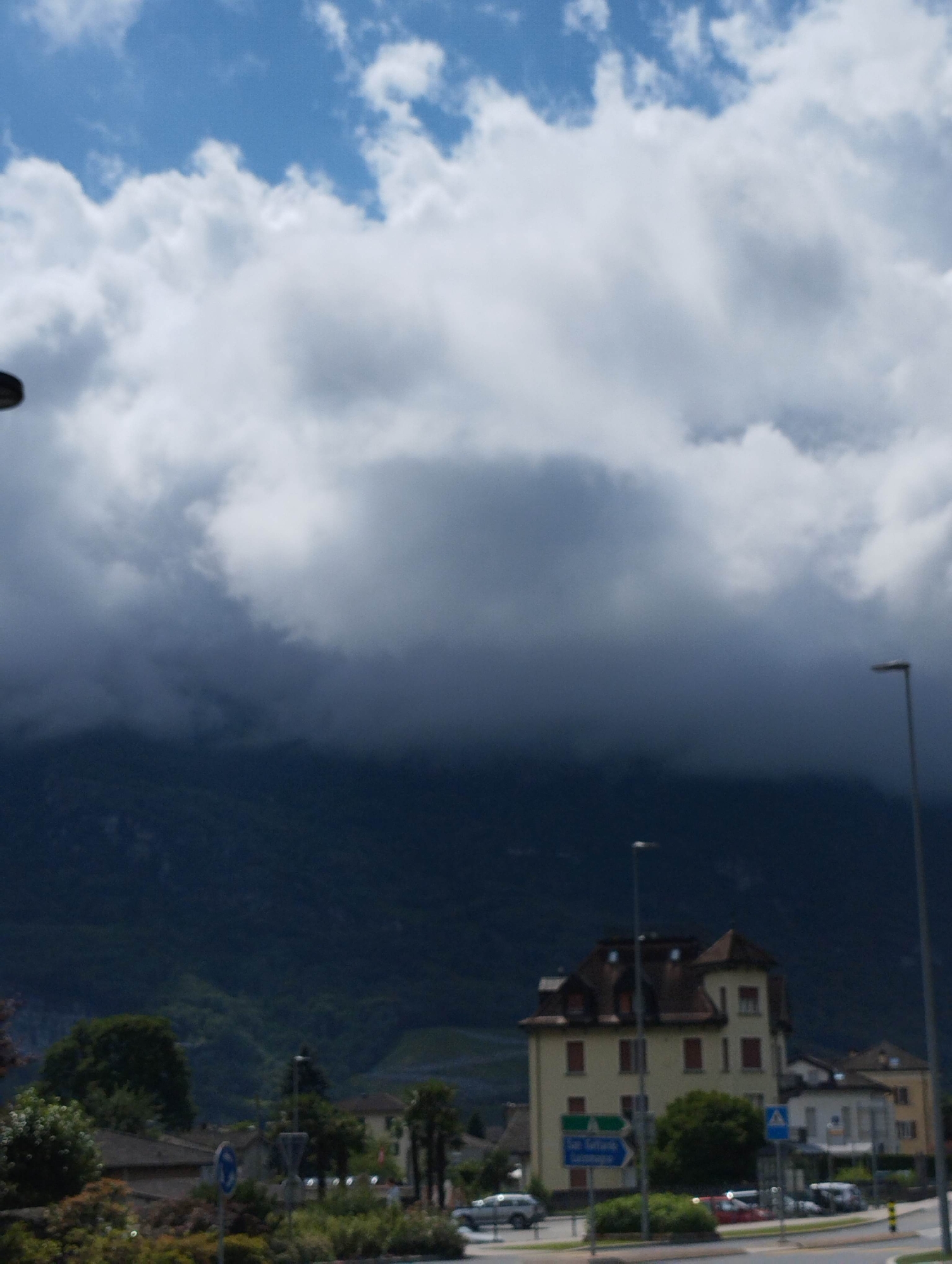 The image captures a landscape scene featuring a blend of natural and urban elements under a cloudy sky. The upper portion of the frame shows a mix of bright white cumulus clouds against patches of light blue sky. These clouds transition into a dense, dark grey mass that descends to obscure the upper and mid-sections of a prominent, dark mountain range, which dominates the background. The lower slopes of the mountains reveal some verdant foliage.
In the mid-ground and foreground, an urban setting is present. A multi-story, cream-colored building with a complex brown roof and numerous red-shuttered windows stands out. Adjacent to it are other residential-style buildings. Several streetlights, including two tall grey poles on the right and a partially visible dark pole on the far left, illuminate the scene. Road signage is visible, including a blue directional sign with illegible white text and a blue circular sign to its left. A road with a visible white SUV on the left and other vehicles further along, winds through the foreground. Abundant green trees and bushes are scattered across the scene, particularly on the left and in front of the buildings. The overall impression is that of a town situated at the base of large, cloud-covered mountains, likely in a temperate region. No individuals are clearly discernible.