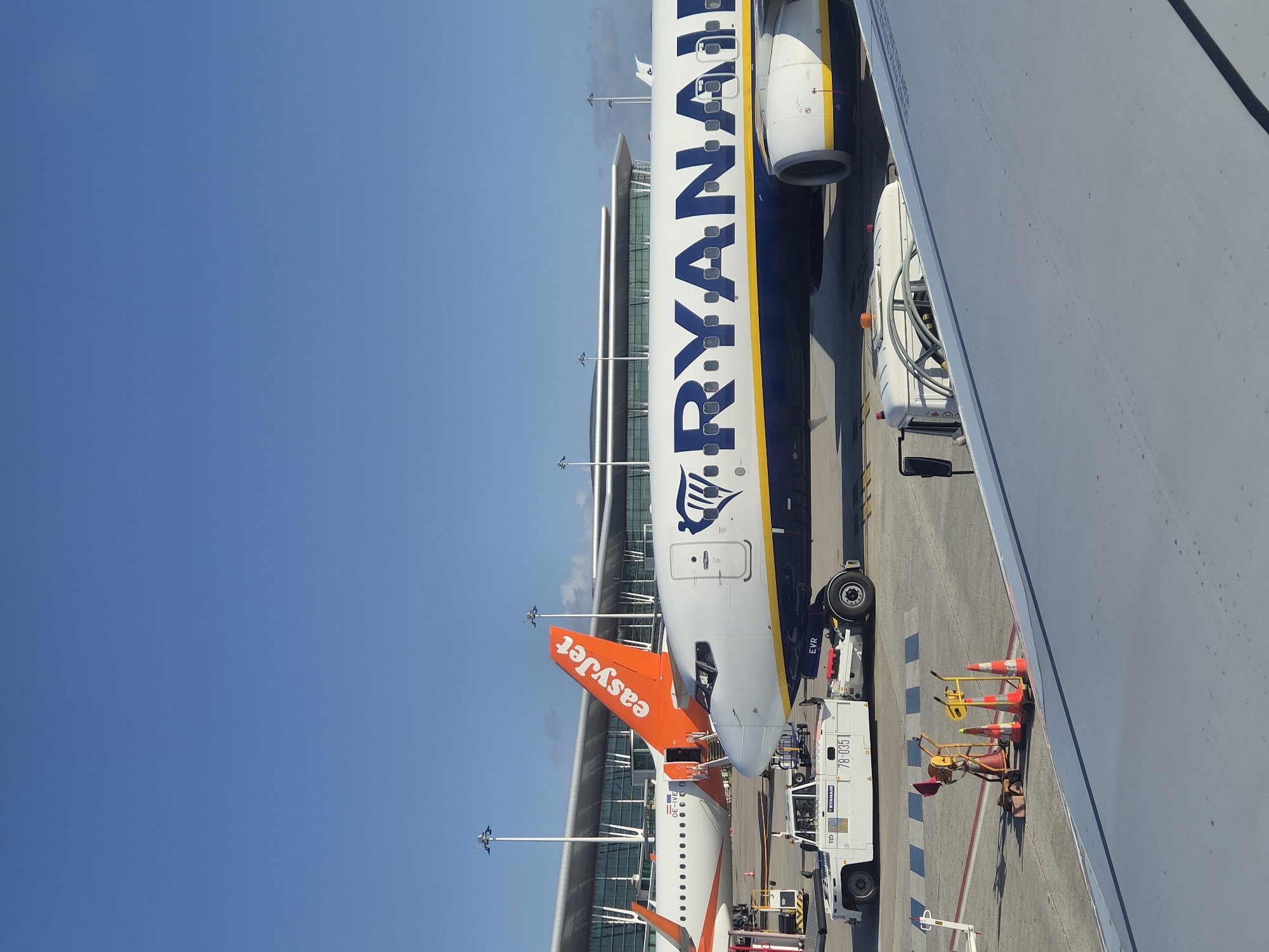 The image depicts a Ryanair aircraft parked at Francisco Sá Carneiro Airport in Moreira, Portugal, adjacent to an EasyJet aircraft. The Ryanair aircraft is white with a blue and yellow stripe along the fuselage and the Ryanair logo. The EasyJet plane is white with an orange tail fin displaying the company logo. The aircrafts are parked on a paved apron next to a modern airport terminal building with a glass facade. Ground service equipment, including tugs, baggage carts and safety cones, surround the planes. The sky is clear and blue. The angle of the photograph suggests it was taken from inside another aircraft, looking down at the other planes.