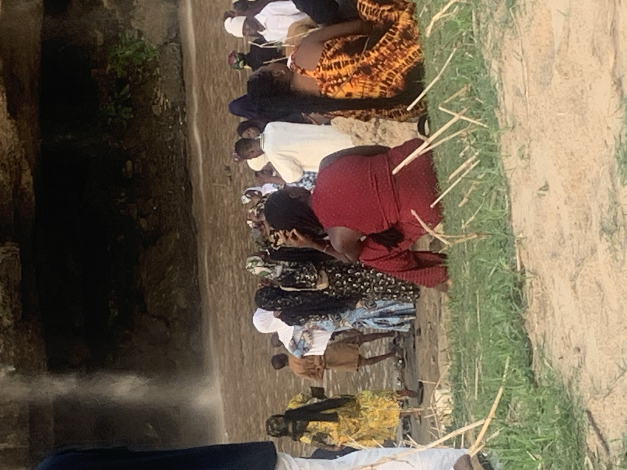 The image depicts a gathering of people at the Gurara Falls in Kukum, Nigeria. A line of approximately 10 individuals, mostly women, are positioned near the edge of the falls. They are kneeling or crouching, possibly in prayer or observation. The environment shows the rocky terrain and vegetation surrounding the waterfall. The waterfall itself is partially visible. People are wearing various attires, some with head coverings. Sticks are laid on the ground next to the kneeling persons.