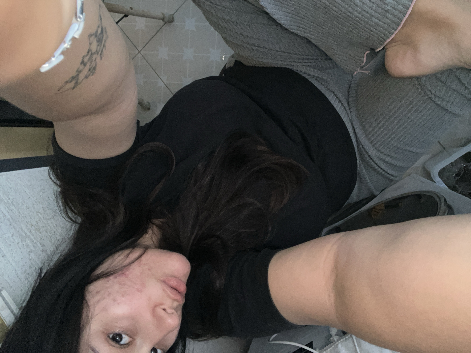 A selfie-style image depicts a person with long, dark hair, wearing a black shirt and gray sweatpants with a pink trim. Their face shows some acne. The person is lying on a light-colored surface, possibly a bed or couch. One arm is extended towards the camera, revealing a tattoo on the upper arm and a silver bracelet. Part of a foot is visible near the top of the frame. The background includes a tiled floor and a glimpse of what may be a chair or table leg. The image does not contain enough information to determine if it was taken in Kiryat Bialik, Israel.