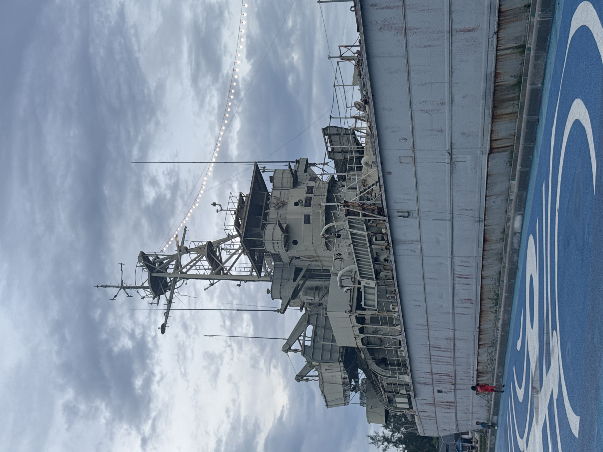The image shows a gray naval ship docked in Thong Sala, Thailand. The ship's structure, with its antennas and communication towers, points toward a cloudy sky. A string of lights is visible above the ship. Along the base, a blue surface with a white design is visible. A person wearing a red shirt is seen walking on the blue surface beside the ship.