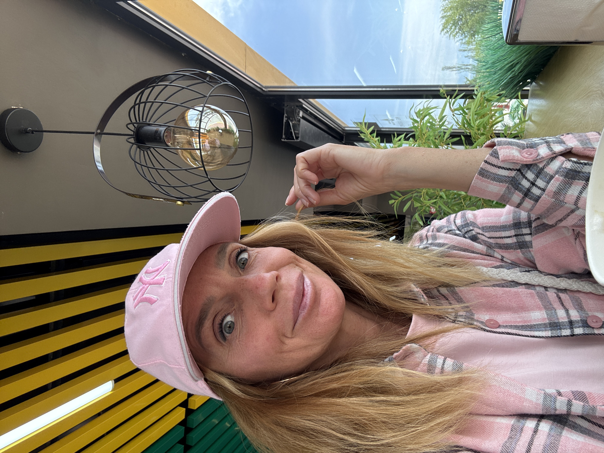 A woman with long blonde hair, wearing a pink baseball cap with a stylized "NY" or "YK" logo, and a pink plaid shirt over a matching pink t-shirt, smiles slightly at the camera. She appears to be seated outdoors or in a semi-outdoor setting, possibly a cafe or restaurant, in Кашат, Kyrgyzstan. Her right hand is raised near her head, lightly touching her hair. In the background, a modern light fixture with a black wire cage and an Edison bulb hangs. Above, a clear sky with scattered clouds is visible through what looks like a glass roof or canopy. To her left, vibrant yellow and black horizontal slat-like structures are visible, transitioning to green slats further down. Lush green foliage is visible through a glass partition behind her, suggesting an open-air or well-lit environment during the daytime.