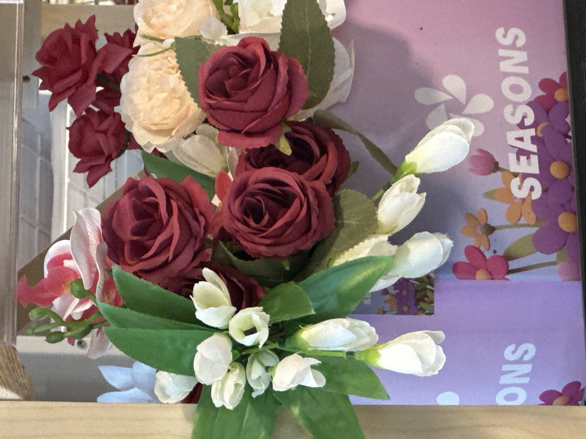 The image shows a floral arrangement of red and peach roses and white buds with green leaves in front of a book titled "Seasons." The flowers appear to be artificial and are arranged on a light-colored surface. The book is lilac-colored. There is no indication of Moscow, Russia.