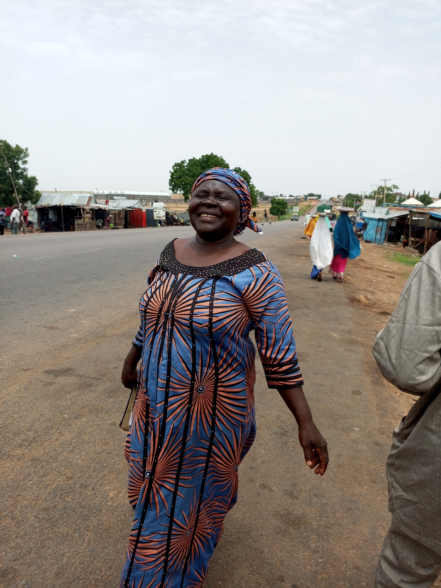 An adult Nigerian woman is pictured standing on a paved road in Gadao, Nigeria, with a light-colored, overcast sky above. She is smiling widely and looking towards the camera. She is wearing a blue and orange patterned dress with a matching headscarf. Her left hand is extended slightly, and she is carrying a bag. In the background, there are several open-air market stalls and buildings lining the road. Other people are visible in the distance, some walking and others gathered around the stalls. The road appears to be a main thoroughfare with vehicles present.