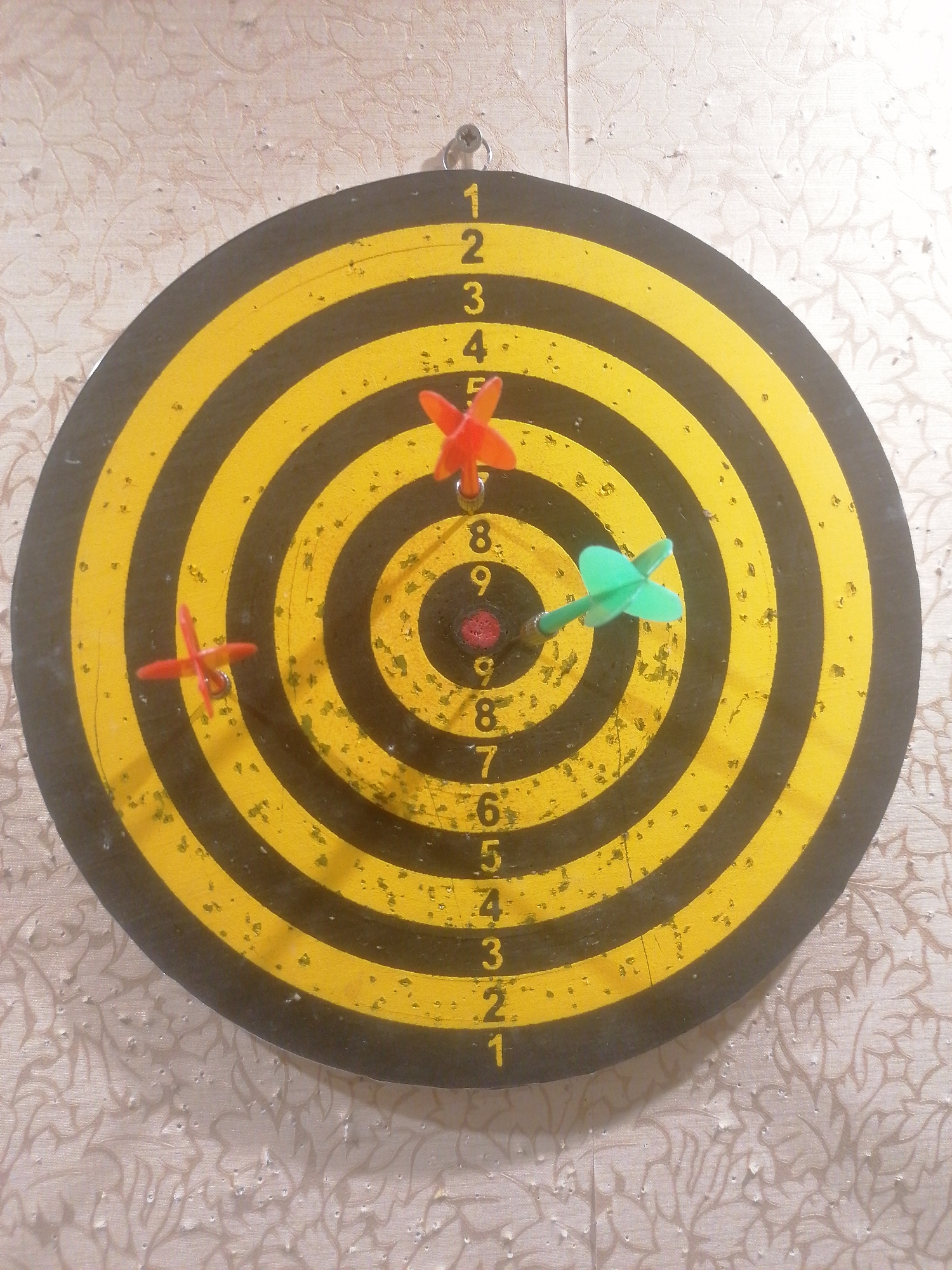 A dartboard with three darts in it is hanging on a wall with a floral pattern. The dartboard is circular with alternating yellow and black rings. Numbers are visible on the left side of the dartboard, ranging from 1 to 9. One red dart is in the yellow ring with the number 4. Another red dart is in the black ring with the number 8. A green dart is in the yellow ring with the number 8. The dartboard appears to be used and shows signs of wear and tear, with small flecks of paint missing. The wall behind the dartboard has a light-colored floral wallpaper with a textured finish. A metal hook is visible at the top of the dartboard, used to hang it on the wall. There are no people or animals in the image. The scene is indoors, in a residential setting. No specific location in Angarsk, Russia can be identified from the image.