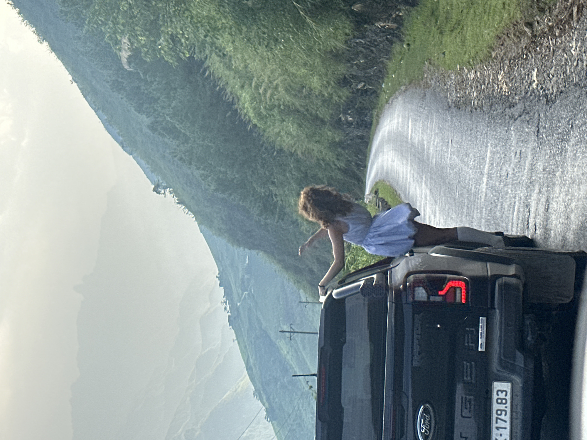 A medium shot depicts a woman partially outside a dark-colored Ford pickup truck traveling on a road in Du Già Commune, Vietnam. The woman is leaning out of the vehicle, facing left with her right arm extended. She wears a light-colored dress and dark-colored footwear with white socks. The truck has a license plate number 30K-179.83. The road is asphalt and bordered by green vegetation. The background showcases a mountainous landscape under an overcast sky.