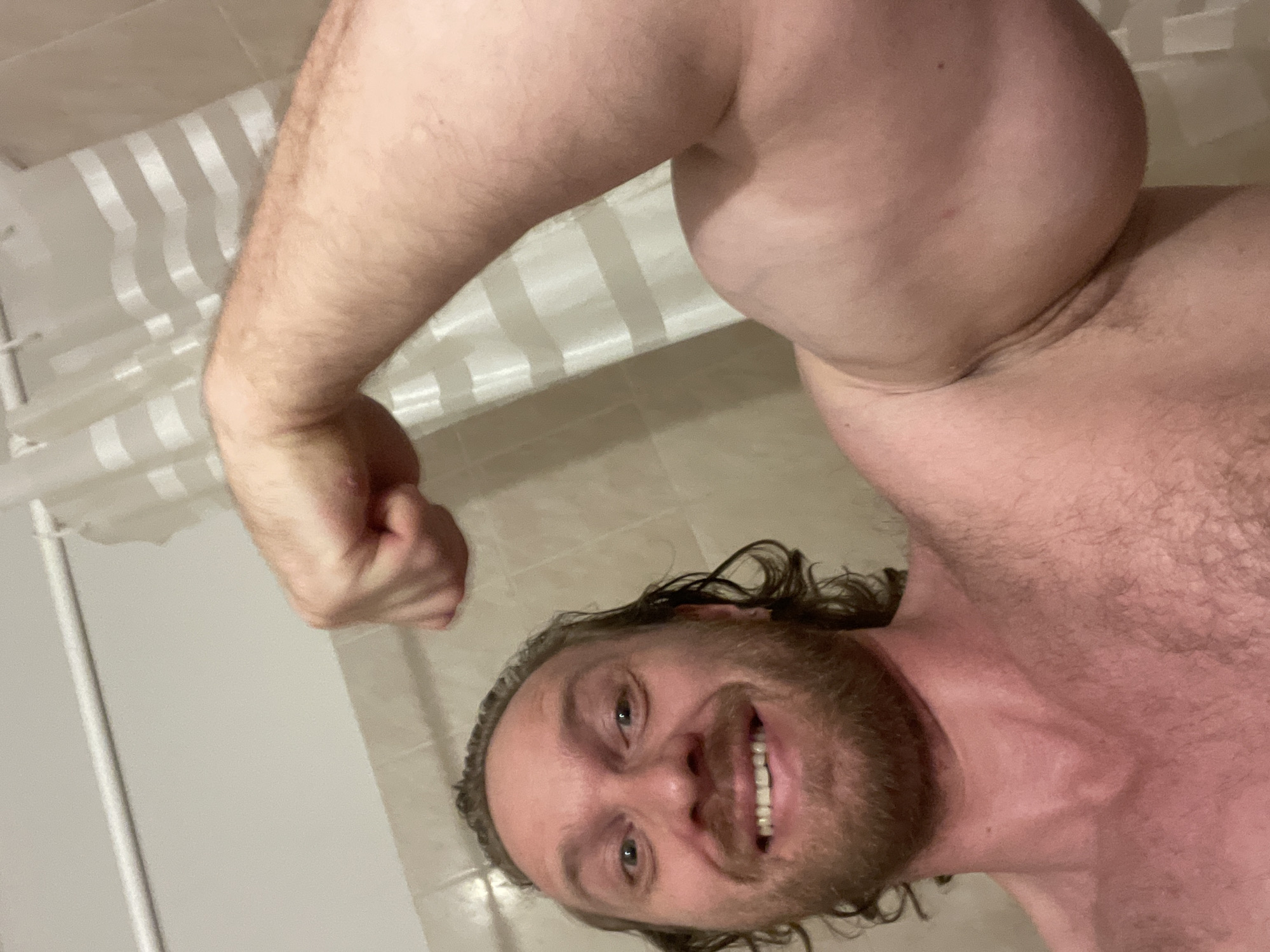 The image shows a man flexing his bicep. He is smiling and looking at the camera. He has medium-length brown hair and a beard. He is wearing a shower curtain as a backdrop. The image is taken indoors, likely in a bathroom. There is no indication of the location in Saint Petersburg, Russia.