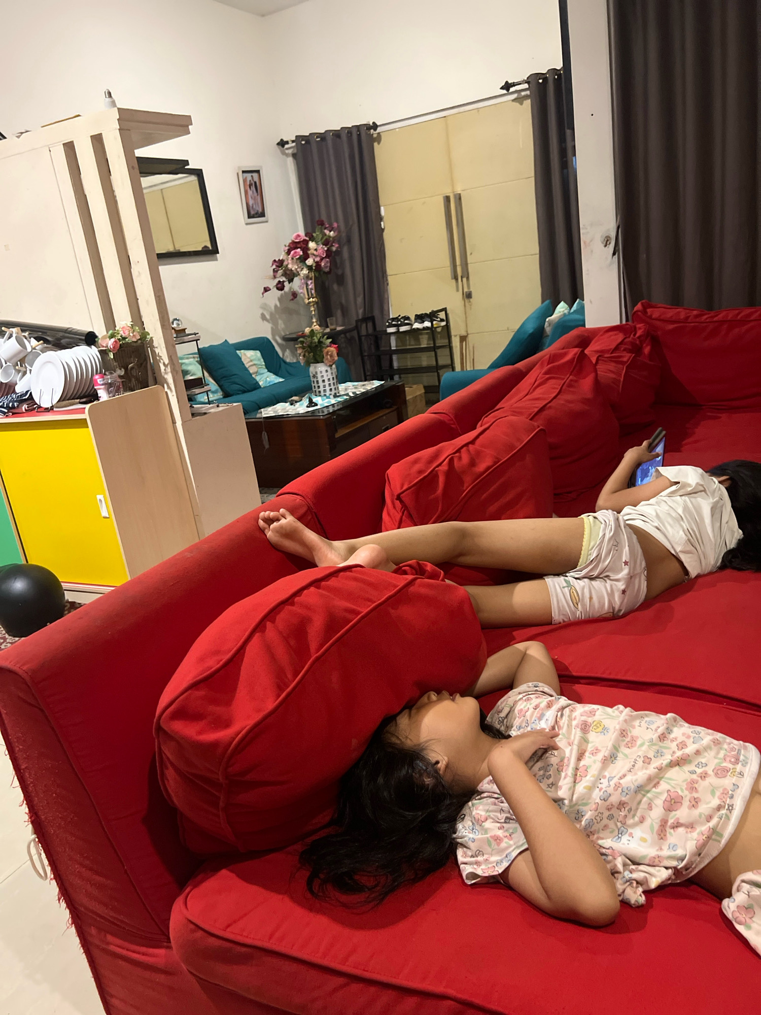 Two young children are lying on a red sectional sofa in a living room, likely in Sleman, Indonesia. The child on the left is lying on their back with their head resting on a red pillow, wearing a light-colored pajamas with a floral print. The child on the right is lying on their side, facing away from the camera, and is looking at a mobile phone. This child is wearing a white t-shirt and light-colored shorts with a fruit print. The sofa is large and appears to be made of fabric. In the background, there are grey curtains, a wooden cabinet with a mirror, and a turquoise sofa. The room is dimly lit, suggesting it is nighttime or the lights are turned down.