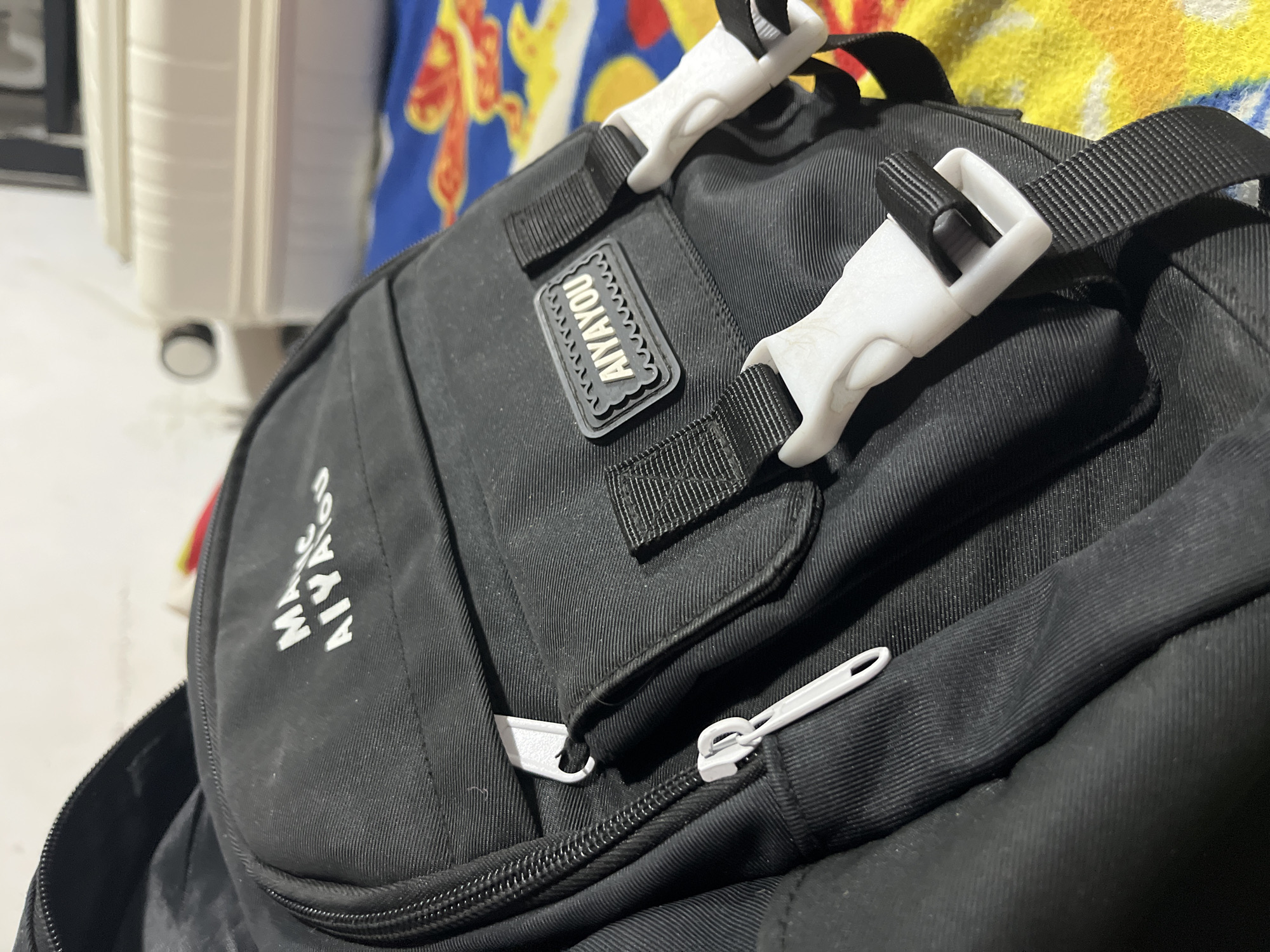 The image shows a close-up view of a black backpack. The backpack features white plastic buckles and a gray rubber patch with the word "AIYAYOU" printed on it. The backpack also has white zipper pulls. In the background, a white suitcase is visible. The setting appears to be indoors. Location information (Juan Griego, Venezuela) is not visually discernible from the provided image.