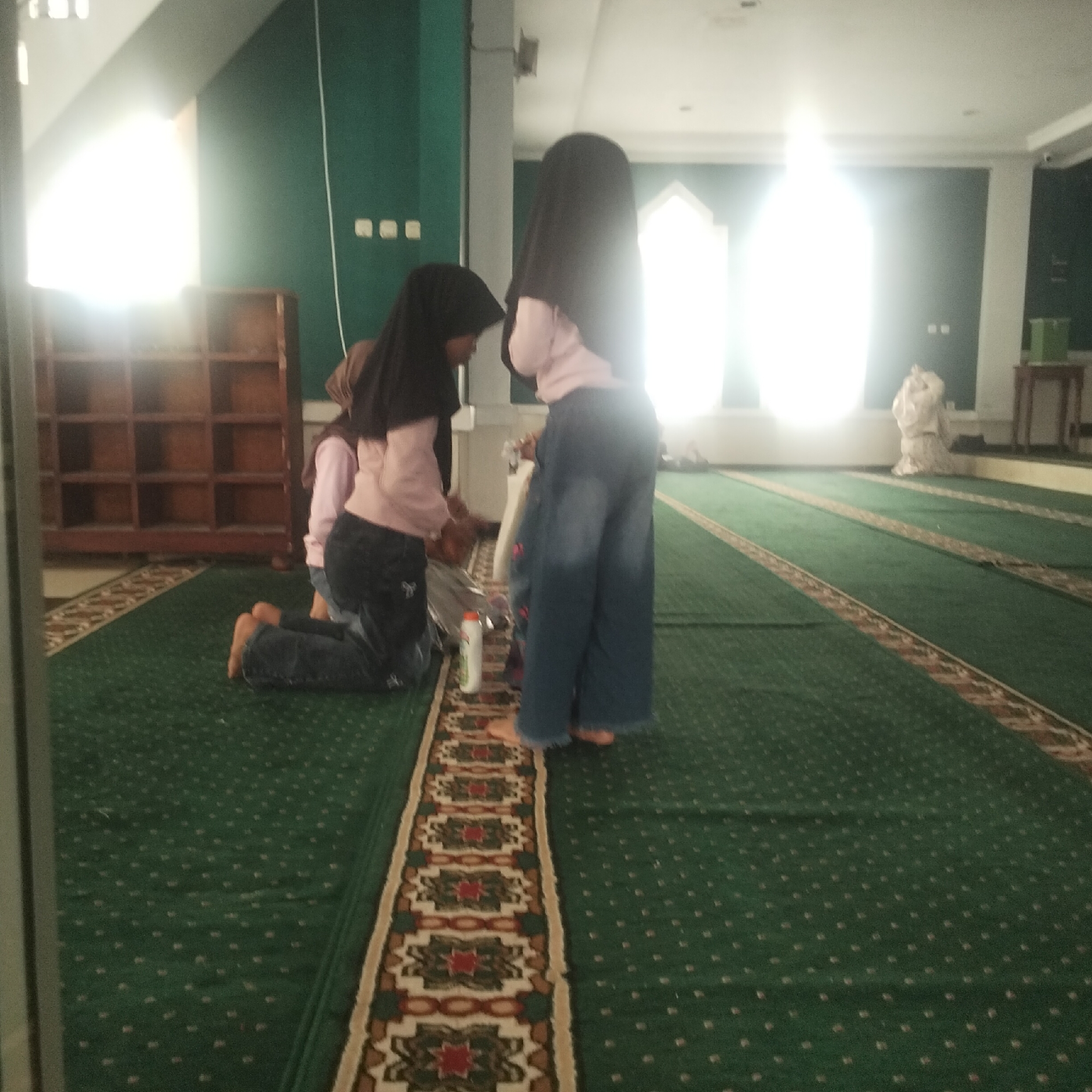 In the city of Cisaat, Indonesia, within what appears to be a mosque, two young girls are present. One girl, wearing a pink shirt and dark headscarf, is kneeling on the green carpet. Another girl, also in a dark headscarf, stands to her right, facing away from the camera. She wears a light-colored shirt and wide-legged denim pants. The room features a green carpet with decorative runners and a wooden shelf unit against the left wall. Bright light emanates from windows on the far wall. The overall scene suggests a moment of prayer or religious observance.