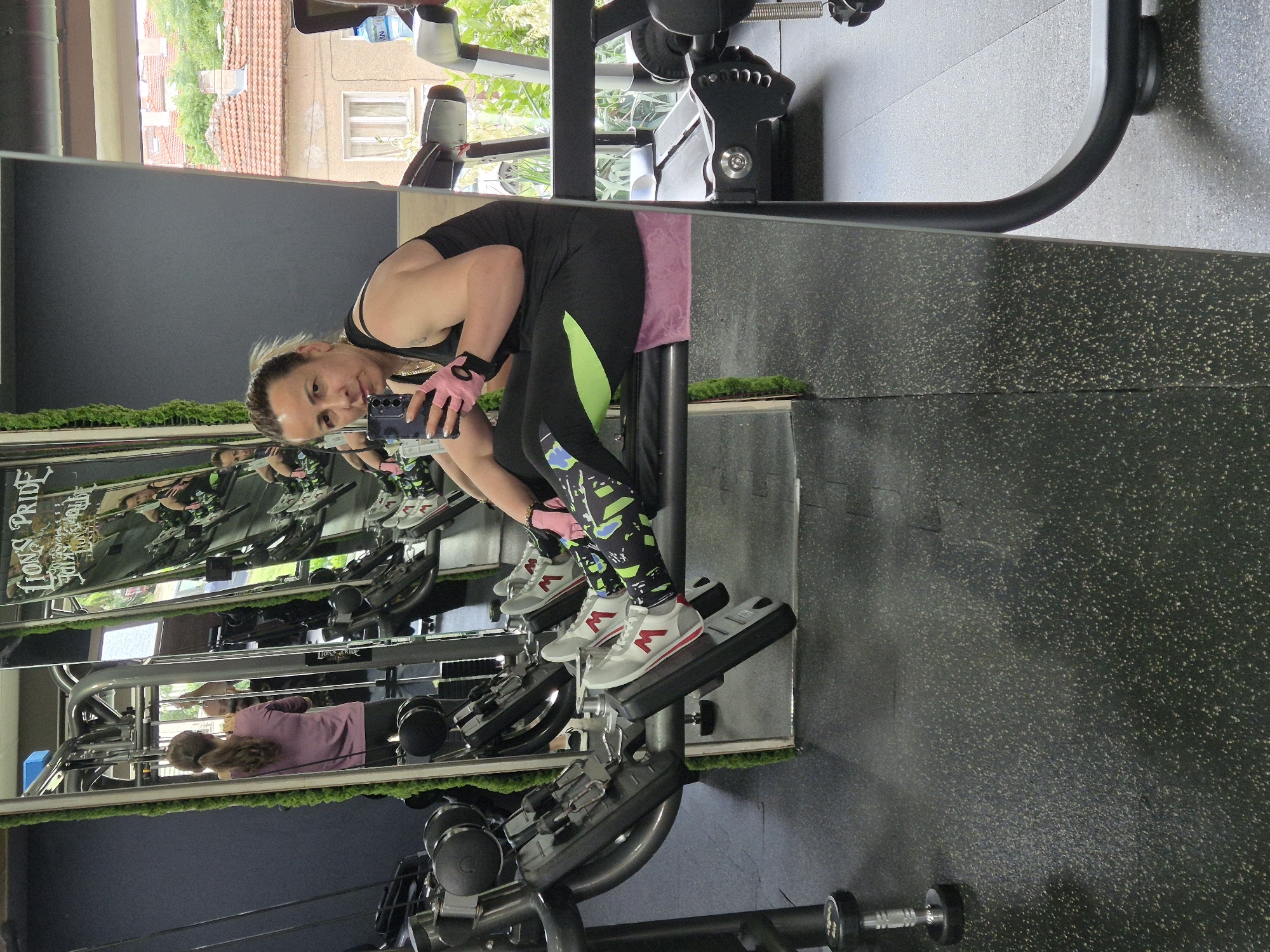 The image shows a woman in a gym, taking a selfie in front of a mirror. She is wearing black workout clothes with neon green accents and patterned leggings, along with pink gloves. She is seated on a leg press machine, holding a smartphone. The gym has a mirror that extends along a wall, and moss has been attached to its perimeter. Reflections in the mirror show other gym equipment and another person in workout clothing. Outside the window, there are residential buildings and greenery. The city and specific location within it are not identifiable.