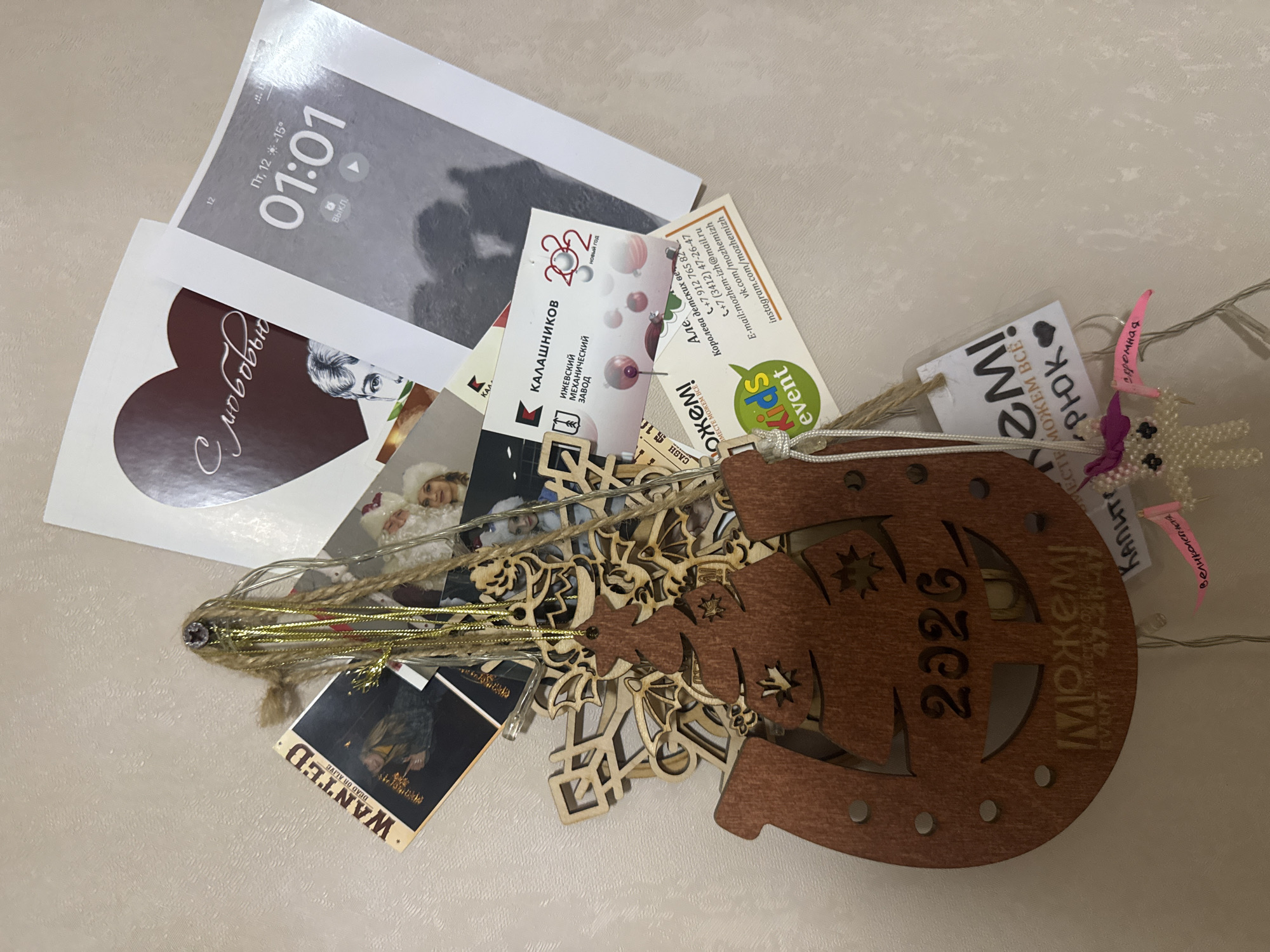 This image captures a collection of miscellaneous items, predominantly printed cards and decorative elements, artfully arranged on a light, textured surface.
The central subject is a large, brown wooden horseshoe-shaped ornament with intricate cut-outs, featuring the year "2026" engraved on it. Adjacent to it are smaller, lighter wooden snowflake-like decorations and a string of delicate fairy lights intertwined with golden tinsel and natural twine.
Beneath and around these decorations are various paper and card items. One prominent card is a printout resembling a phone screen, displaying "01:01" as the time, "Пт, 12 - +15°" (Friday, 12th - +15°C) for date and temperature, and "Выкл." (Off). Another heart-shaped card is visible, bearing the text "С любовью" (With love).
Business cards and small tags are also present. One business card pertains to "КАЛАШНИКОВ" (Kalashnikov), specifically the "ИЖЕВСКИЙ МЕХАНИЧЕСКИЙ ЗАВОД" (Izhevsk Mechanical Plant), adorned with "2022 новый год" (2022 New Year) and festive ornaments. Another is for a "Kids event," listing contact details including phone numbers (+7 912 765 82, +7 (3412) 47-26-47), email, and social media handles (vk.com/mozhemizh, instagram.com/mozhemizh). This card also features "Можем" (We can) and "Королева детских веч" (Queen of children's evenings).
Other notable prints include a "WANTED DEAD OR ALIVE" poster with an image of a person, and a card showing "CASH $16" next to a person's face. Small white tags are also visible, bearing handwritten or printed Russian words such as "МОЖЕМ ВСЁ" (We can do everything), "РЮК", "скромная" (modest), "Великолепная" (magnificent), and "Капите" (Capite).
The arrangement suggests a collection of personal mementos, business contacts, and festive decorations, possibly accumulated from various events or celebrations over several years, indicated by the "2022" and "2026" dates. No direct activity is shown, but the items themselves tell a story of past interactions and festive occasions.