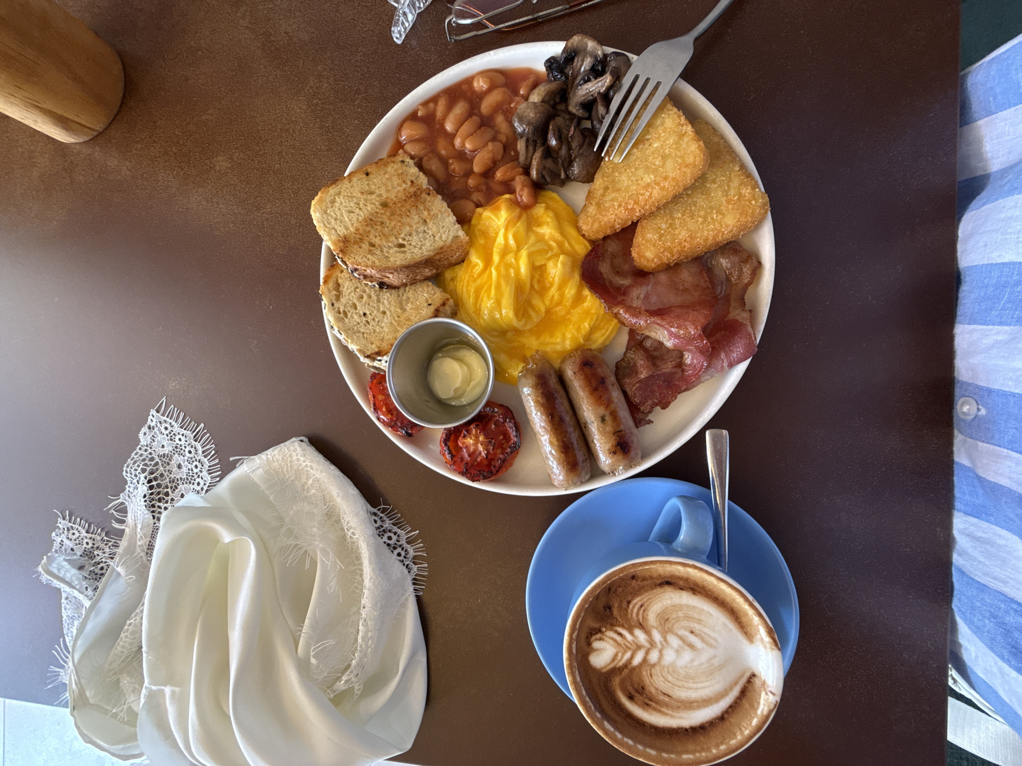 A top-down view shows a hearty breakfast on a dark brown table. The meal is served on a white plate and includes scrambled eggs, two sausages, bacon, grilled tomatoes, baked beans, mushrooms, hash browns, and two slices of toast. A small metal cup holds what appears to be butter or sauce. Beside the plate, a blue saucer holds a latte with intricate foam art. A silver spoon rests in the cup. To the left of the breakfast, a pile of white fabric, possibly a tablecloth or napkin, is draped. In the upper left corner, a wooden object, possibly part of a salt or pepper grinder, is visible. The scene suggests a meal at a cafe or restaurant. The lighting is bright, indicating daytime. A blue and white striped fabric, likely clothing, is visible on the right side of the frame. No visible text. The setting is Kuta, Indonesia, suggesting a tropical, potentially humid environment.