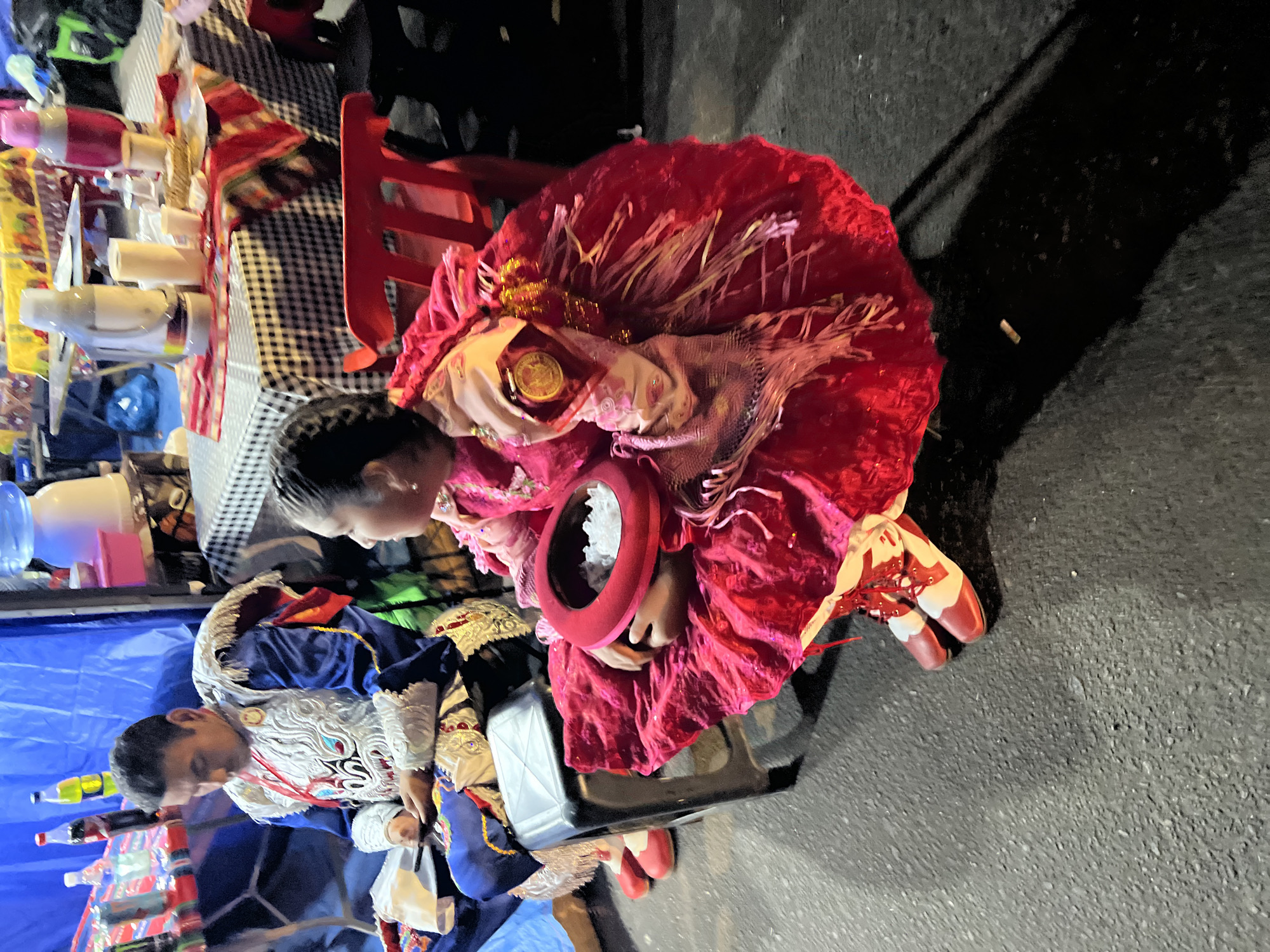 A man in a blue and white traditional costume sits on a black stool. He is looking down, with his left hand holding a phone or small object. To his right, a young girl in a vibrant red dress and white and red shoes kneels on the ground. She holds a red circular object filled with white material. The scene takes place on an asphalt surface, likely a street or public area. In the background, there are tables with various items, including drinks and food, suggesting an outdoor market or event. A blue tarp is visible behind the man. The overall lighting suggests it is evening or nighttime. This scene is likely from the city of Arica, Chile, given the context provided.