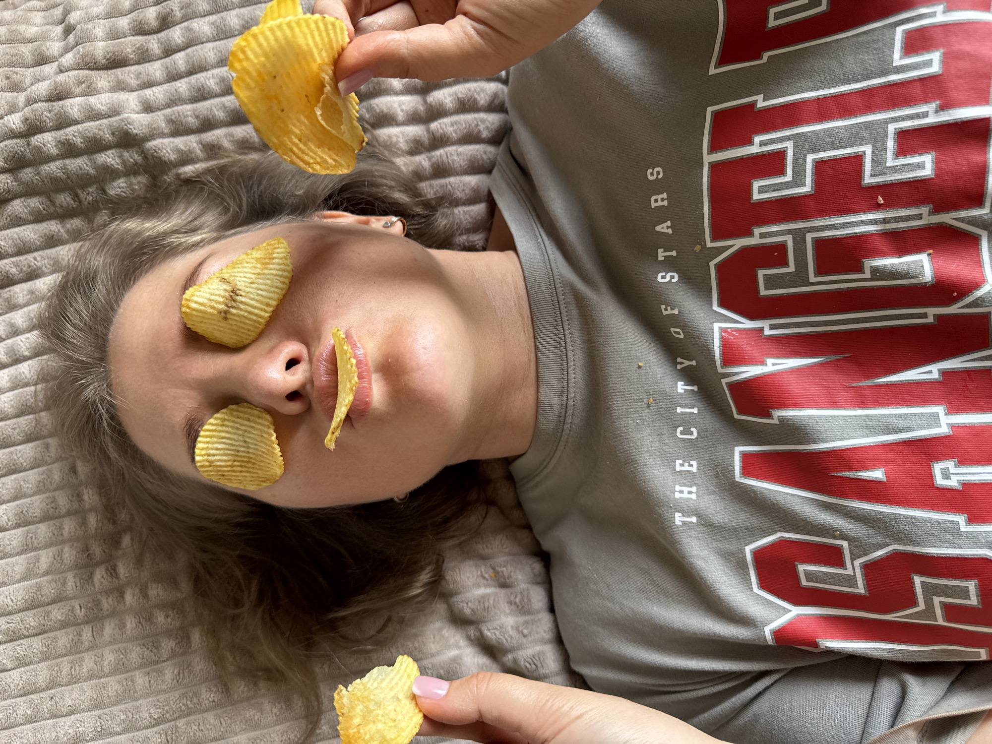 A person lies on a beige, textured surface, likely a bed or blanket, wearing a grey t-shirt with large red letters and the phrase "THE CITY OF STARS" printed vertically in white. The person's face is visible; their eyes are covered with ridged potato chips, and another chip is placed on their upper lip. Two hands are visible, each holding a potato chip above the person's face. The scene appears to be indoors. The location, Ферма, Russia, is not visibly discernible in the image.