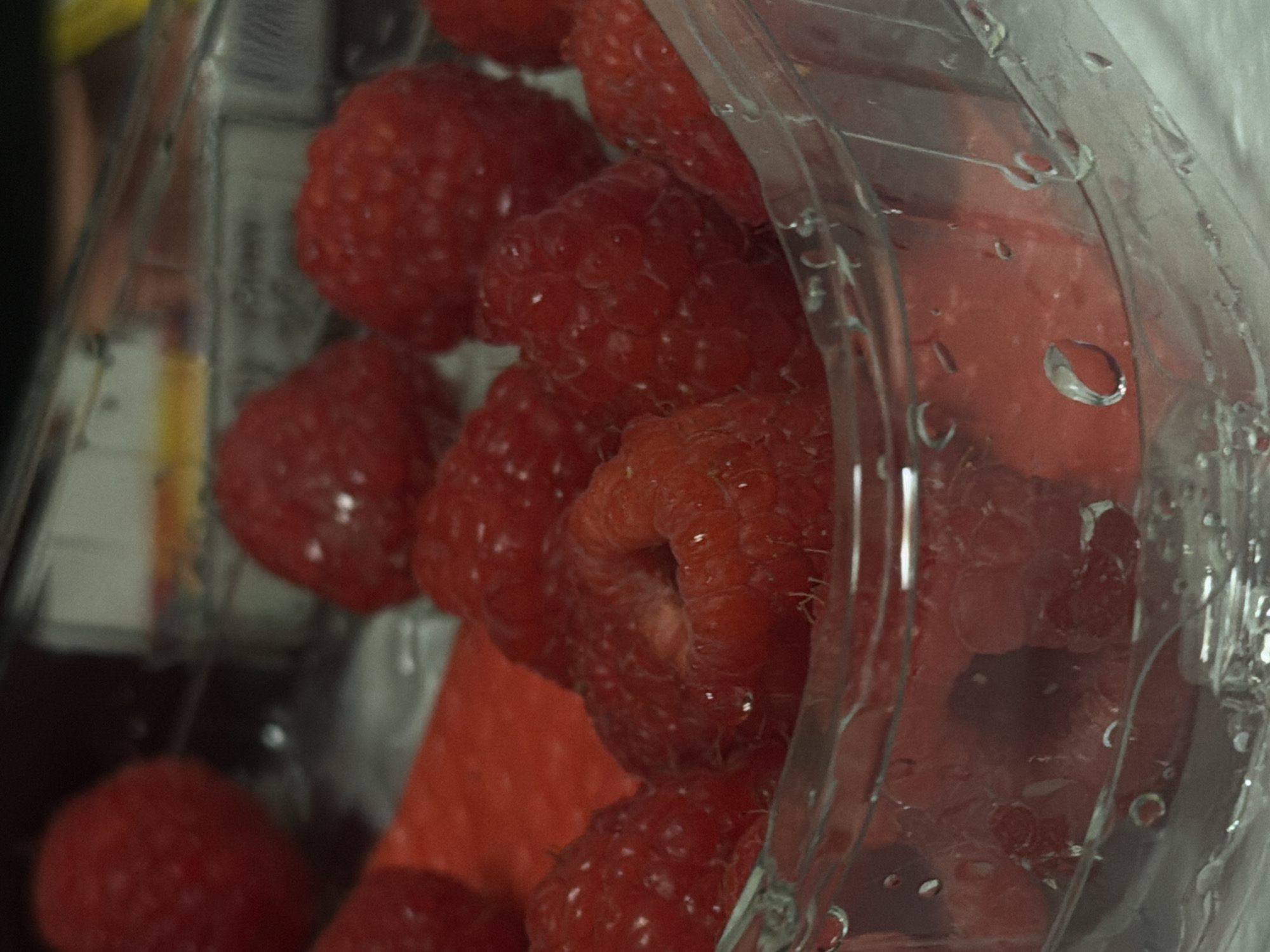 A close-up, eye-level shot features a clear plastic container filled with fresh raspberries. The raspberries are a vibrant red and are clustered together, some appearing plump and others slightly shriveled. The container is positioned slightly off-center, with its transparent walls allowing a clear view of the fruit. Water droplets are visible on the inside and outside of the container, suggesting condensation or recent washing. In the background, out-of-focus elements hint at a shelf or surface, possibly in a grocery store or kitchen setting. The lighting is soft, emphasizing the texture and color of the raspberries.