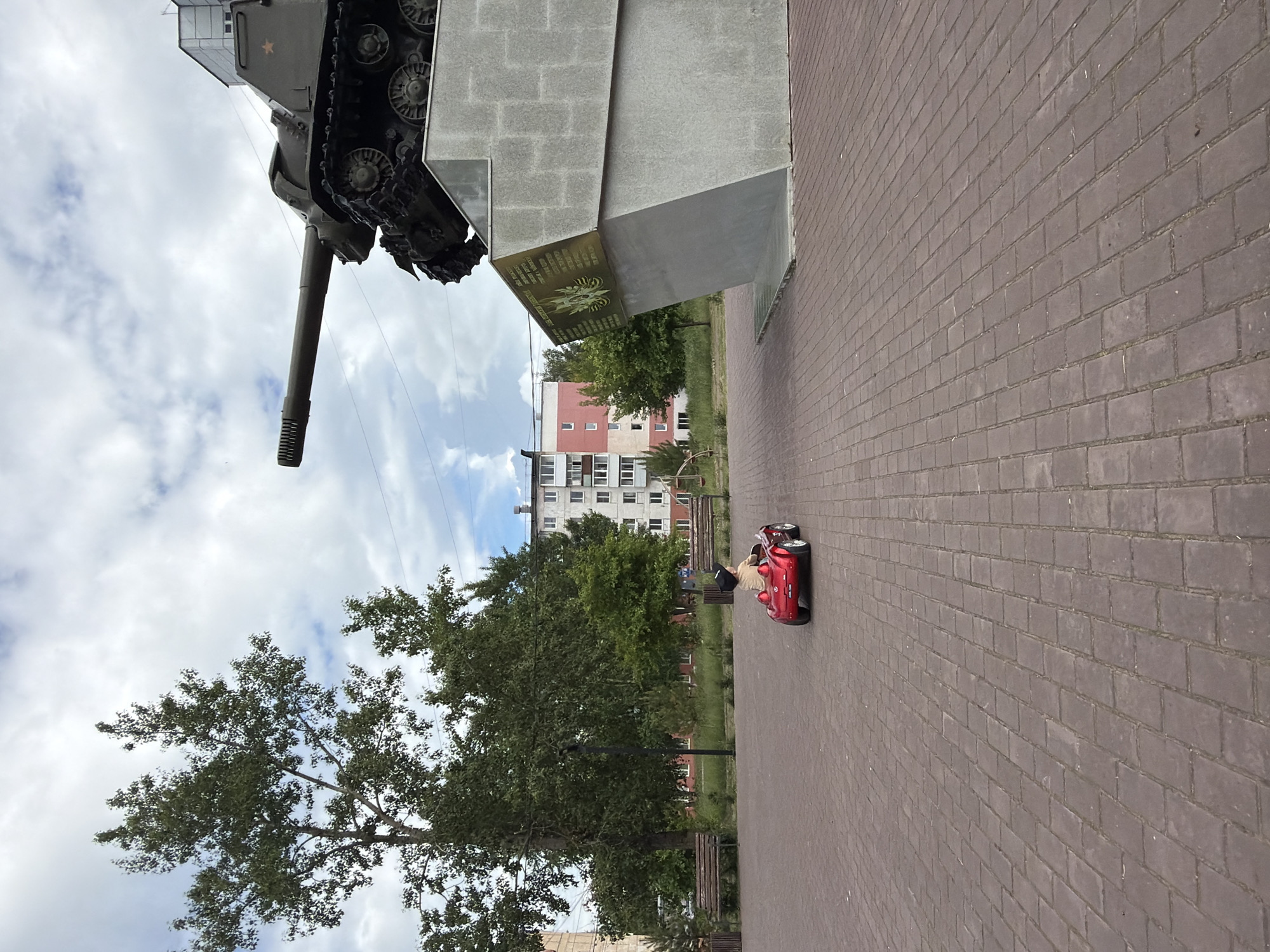 A child is riding a red toy car on a paved plaza in Chaikovsky, Russia. The child is facing away from the camera and is wearing a light-colored shirt and a dark cap. In the background, there is a monument with a tank on top, surrounded by trees and apartment buildings. The sky is cloudy.