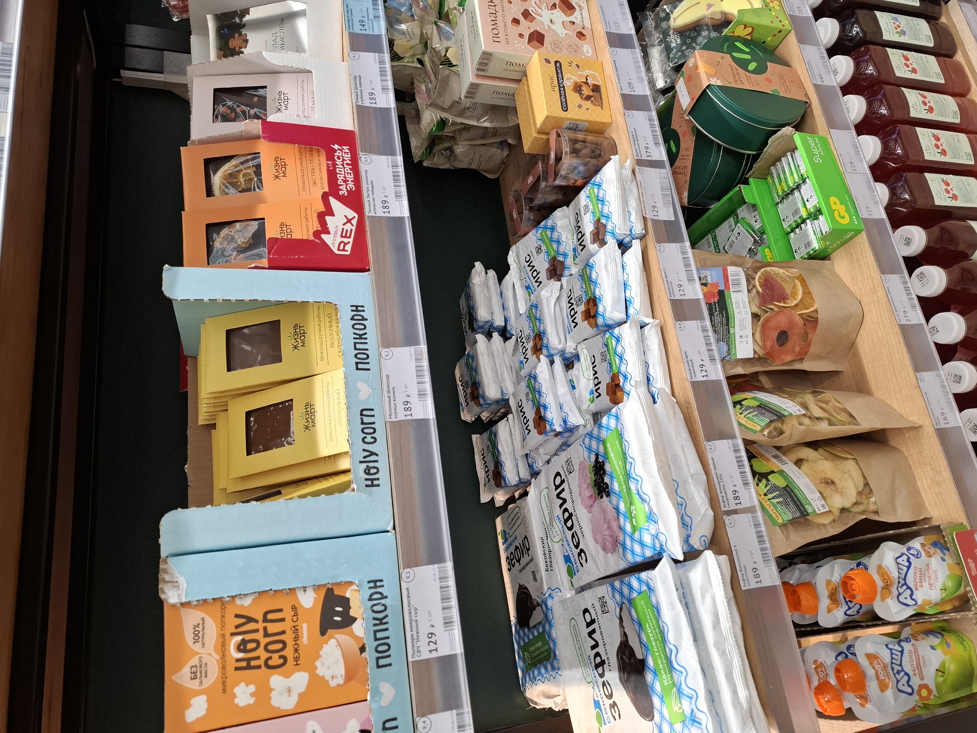 The image displays a grocery store shelf in Tyumen, Russia, stocked with various food products. The items are organized on wooden shelves and include popcorn, chocolate bars, marshmallows, dried fruit, batteries, and bottled beverages. The lighting suggests an indoor environment, typical of a supermarket. No people are visible in the frame. The products are predominantly packaged in colorful wrappers and boxes, with Cyrillic script indicating their origins and contents. Some price tags are visible, providing pricing information for select items.
