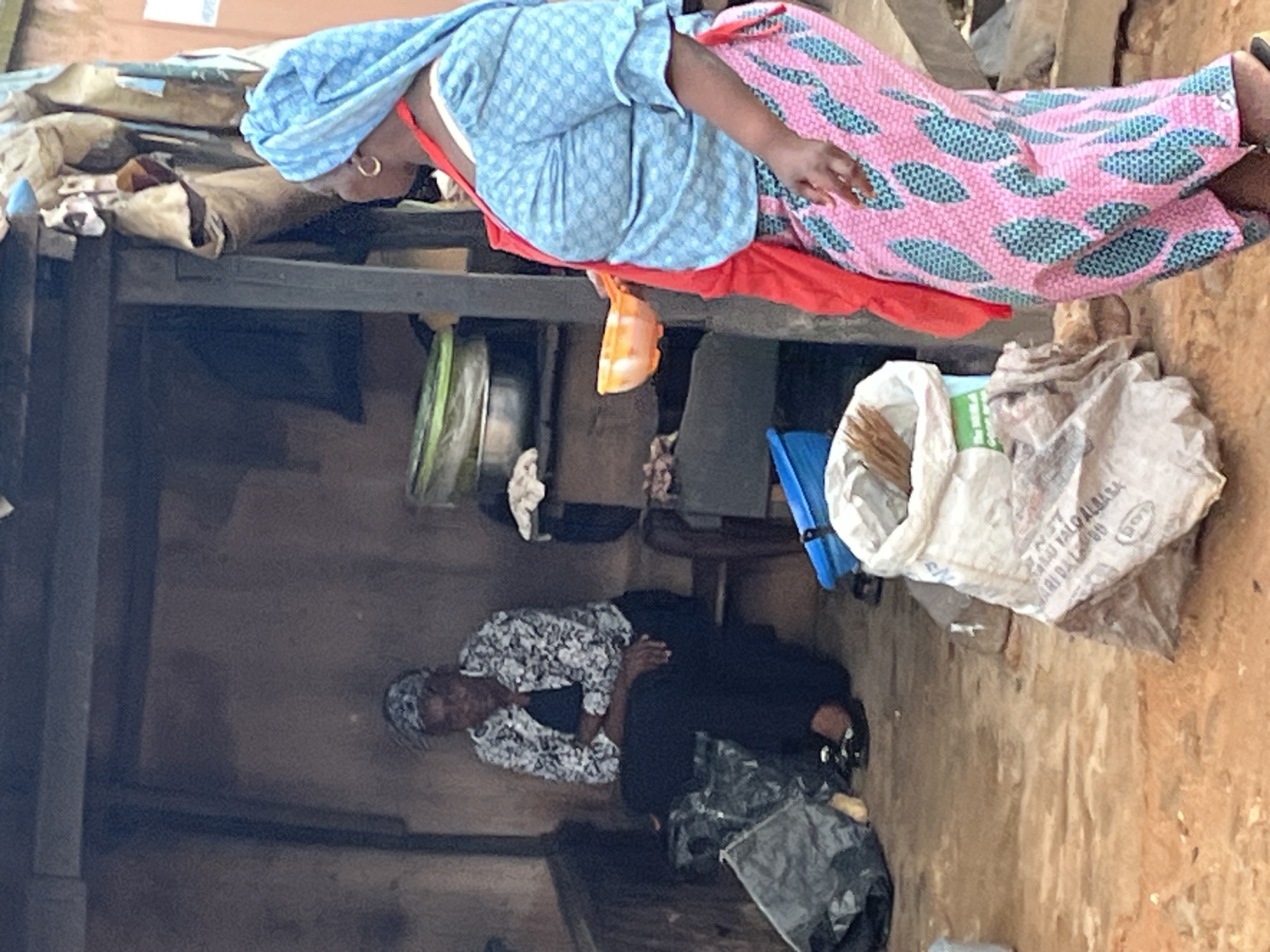 In Jos, Nigeria, two women are seen at an outdoor market stall. One woman is seated on a stool, wearing a black dress and a patterned jacket with a headscarf. She has her arms crossed and appears to be looking down. Beside her are two large black trash bags. The second woman stands in the foreground, facing away from the camera, and is wearing a blue patterned blouse, a pink and green patterned skirt, and a blue headscarf. She is holding an orange bowl with her right hand and has a red cloth draped over her left shoulder. Behind her, a white sack filled with what appears to be dry goods or produce is visible. The stall itself is open-air with a rudimentary structure made of wood and what looks like corrugated metal sheeting. The ground is a mix of reddish dirt and concrete. Various other items, including containers and more sacks, are scattered around the stall.