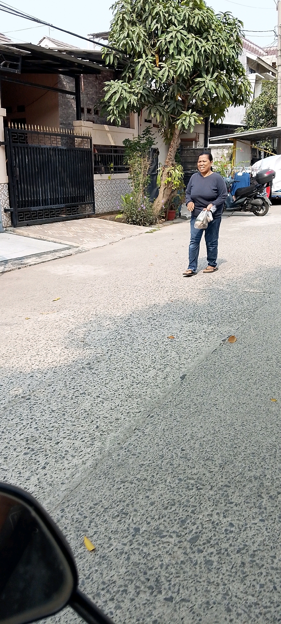 A medium-shot, outdoor, eye-level, full-body, street-level photograph captures a woman walking down a paved street in what appears to be Satriamekar, Indonesia. The woman, who is of Southeast Asian descent, is wearing a dark long-sleeved shirt and blue jeans, with brown flip-flops. She is carrying a transparent plastic bag in her right hand, filled with what seem to be various items. Her facial expression is somewhat strained or pensive, and she is looking slightly off-camera to her left.
The street is paved with a textured asphalt material, with a light-colored concrete ramp leading up to a black wrought-iron gate on the left side of the frame. Behind the gate, a building with a stone-patterned facade and a dark metal overhang is visible. To the right of the gate and the building, a large tree with dense green foliage stands prominently. Further to the right, a motorcycle is parked on the side of the street. The overall environment suggests a residential or mixed-use urban area. The lighting indicates it is daytime, with shadows cast across the street, suggesting a bright, possibly sunny, day. The lower left corner of the image shows a portion of what could be a motorcycle's side mirror, implying the photo was taken from a moving vehicle.